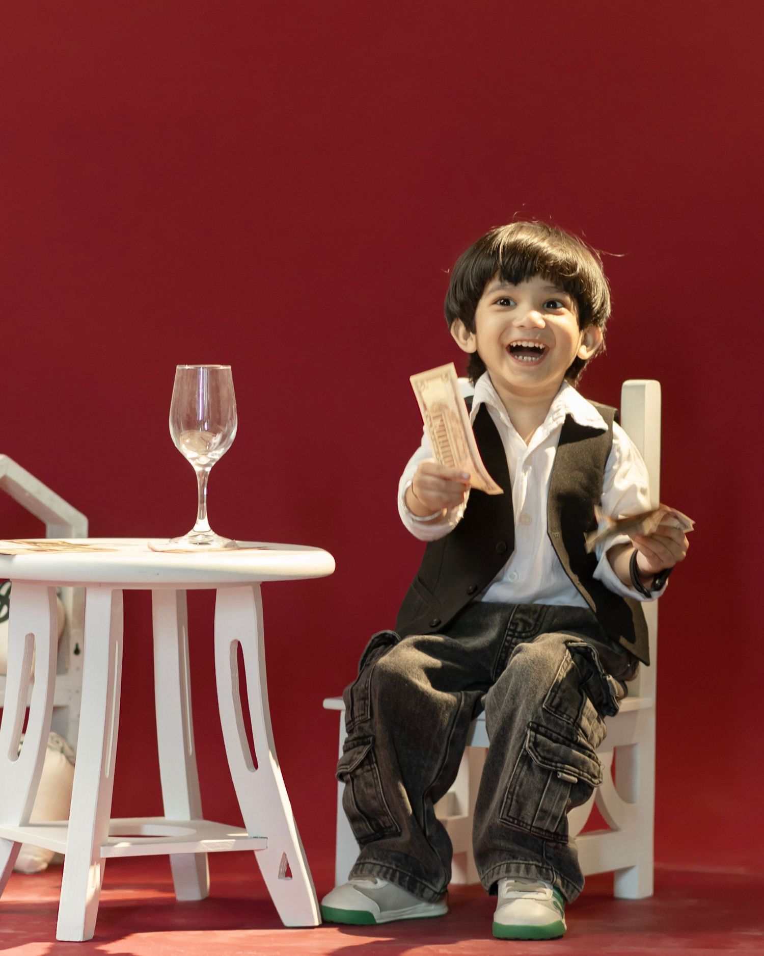 A cheerful young boy in a black blazer sits on a white chair, holding a snack beside a small table with a wine glass.