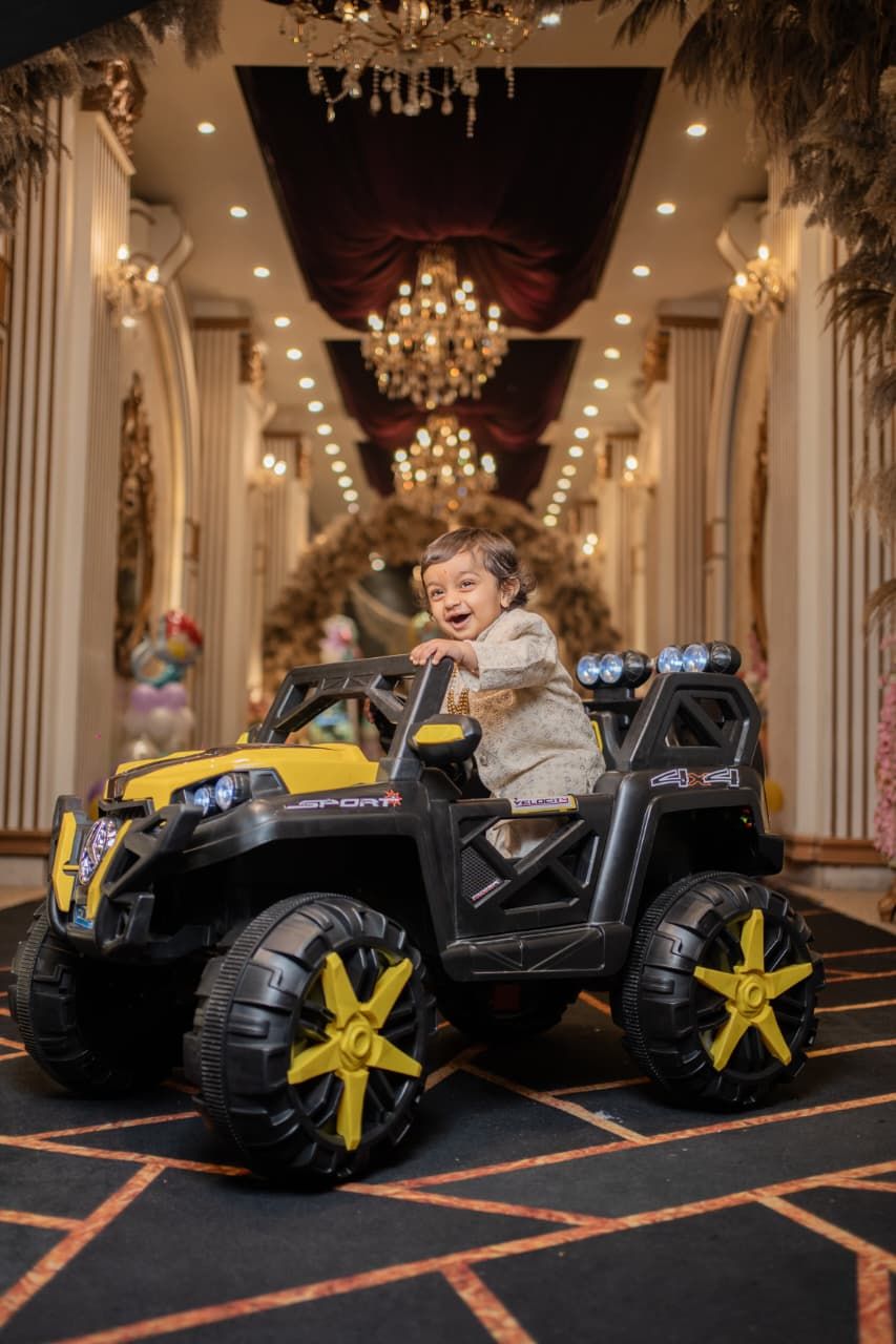 Smiling toddler riding a black and yellow electric toy SUV in an elegant decorated hall with chandeliers.