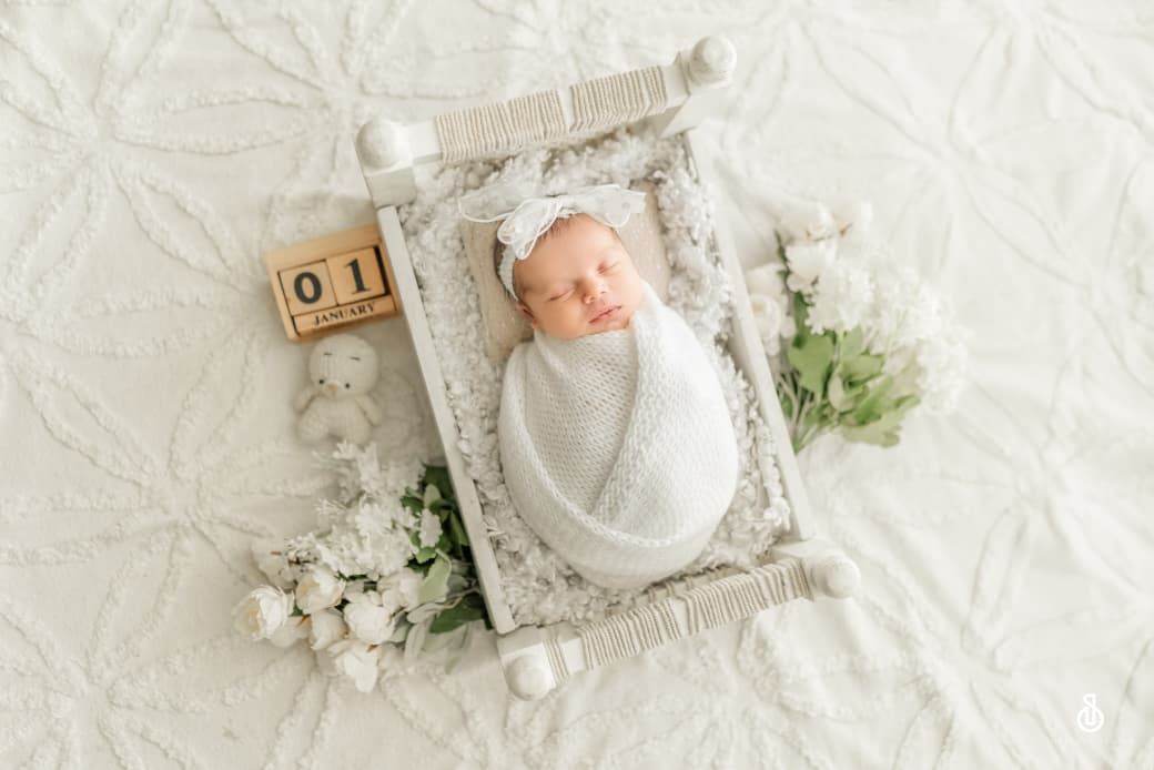 Newborn baby swaddled in white on a tiny bed with flowers and a date block showing 01 in a professional photo.