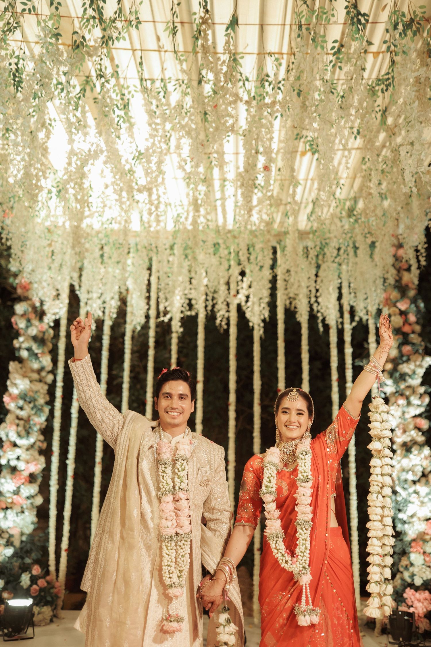 Indian bride and groom in traditional attire celebrate wedding under floral canopy with white hanging flowers.