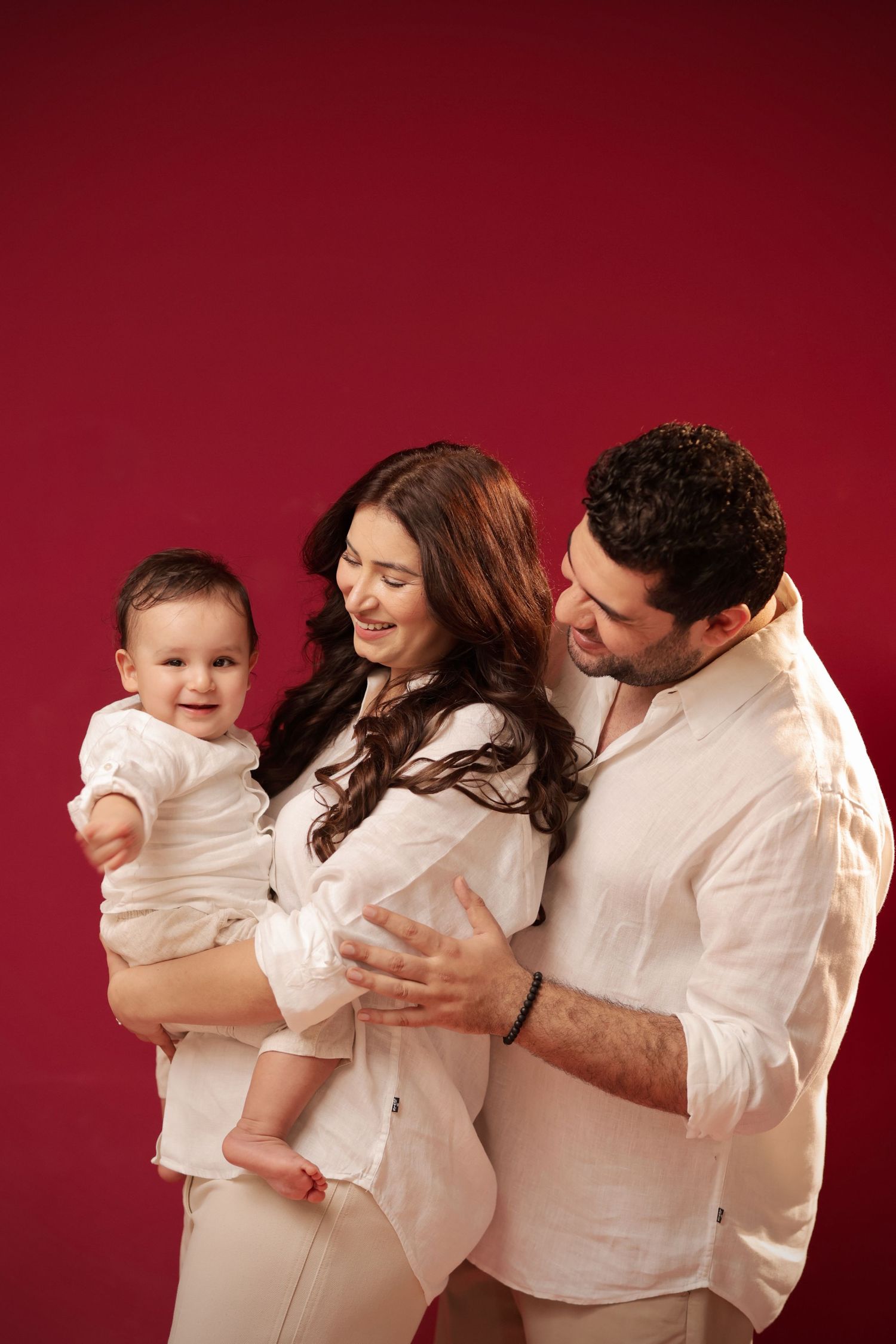 Happy family portrait with parents in white holding smiling baby against red background.