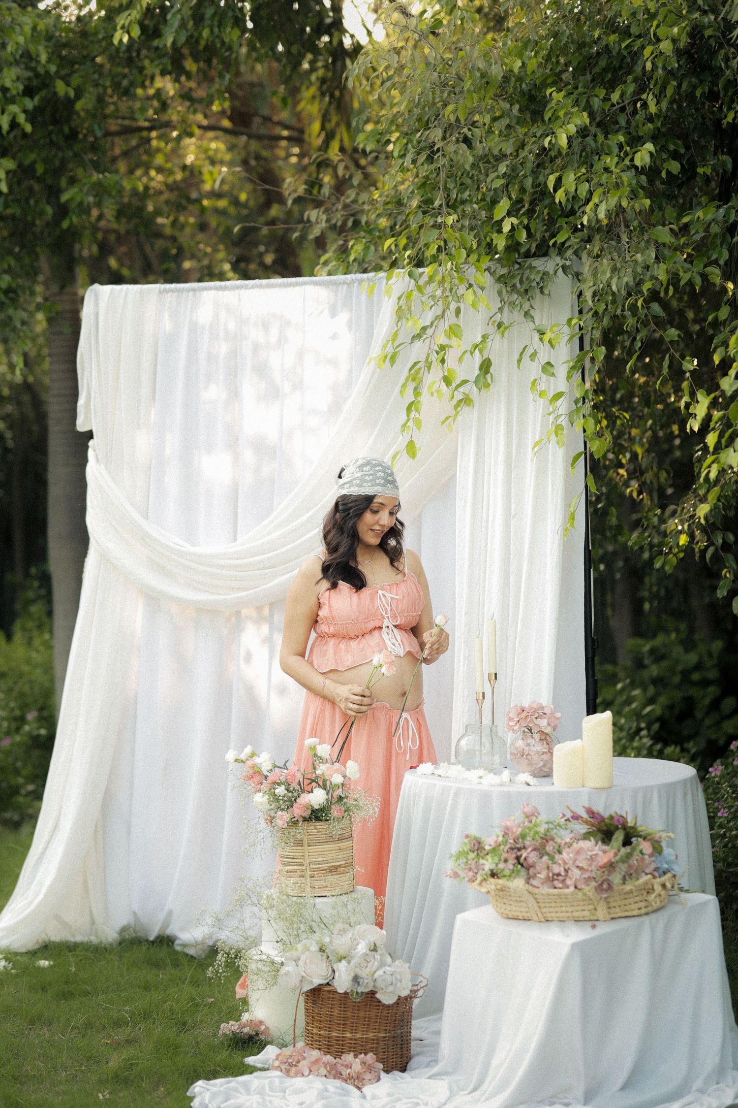 Pregnant woman in pink dress stands by white drape backdrop with flowers at outdoor baby shower.