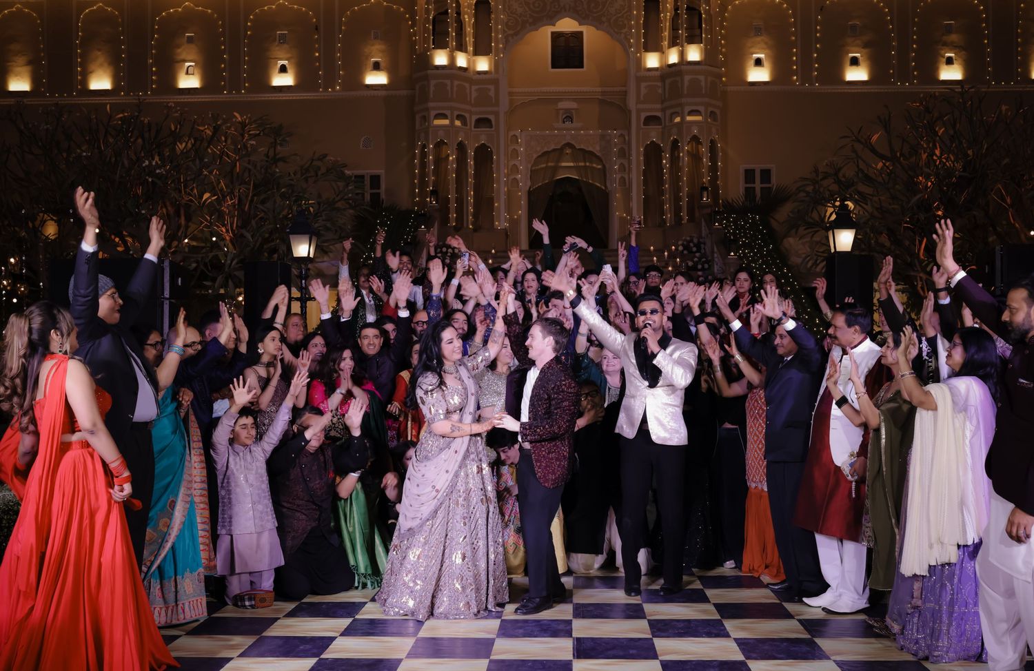 A grand Indian wedding celebration with guests dancing in elegant attire at an ornate palace venue.