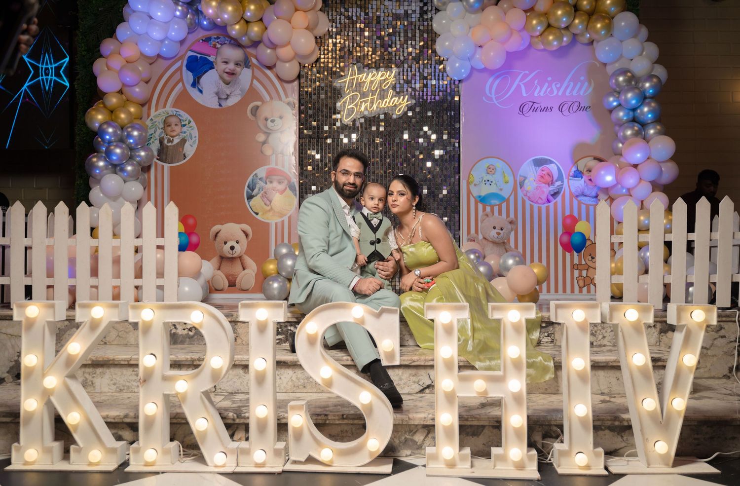 Family posing at Krishiv birthday party with balloon arch, teddy bears, and illuminated name letters on decorated stage.