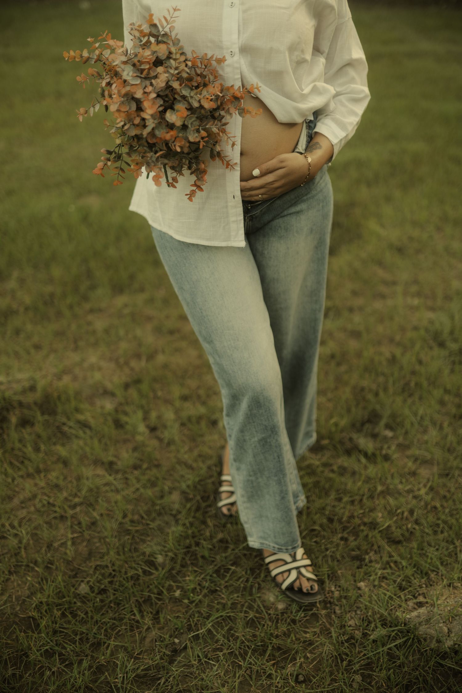 Pregnant woman in white shirt and jeans holding orange wildflower bouquet while standing in a green grass field.