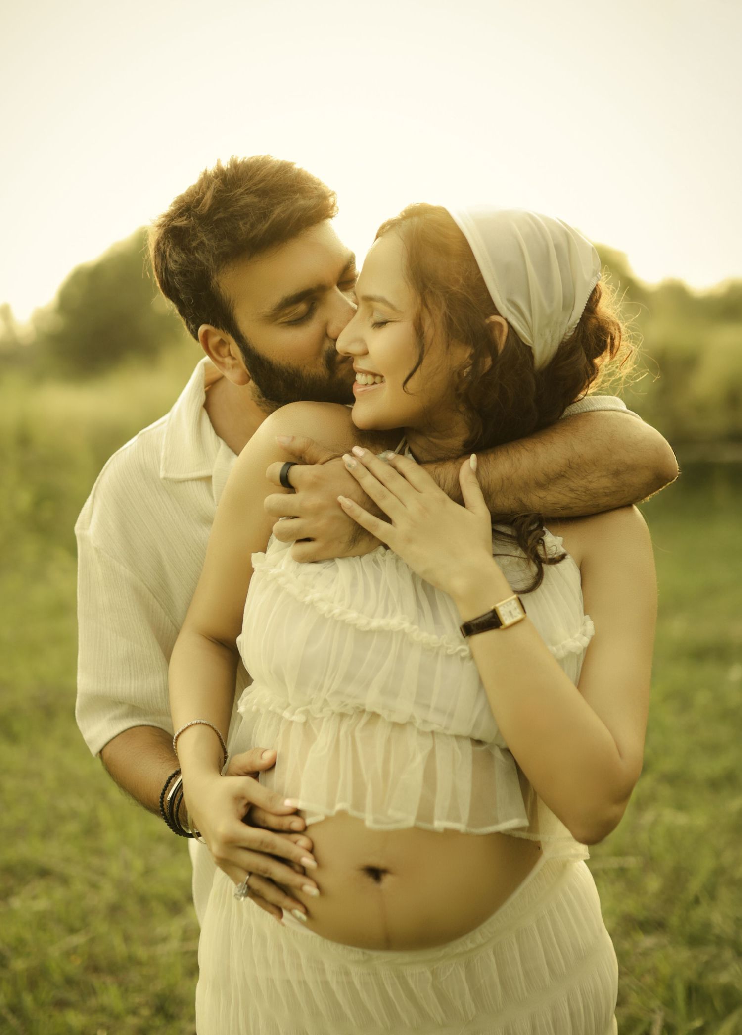 A couple shares a tender embrace outdoors, with the man kissing the woman's head as she smiles, showcasing a maternity moment.