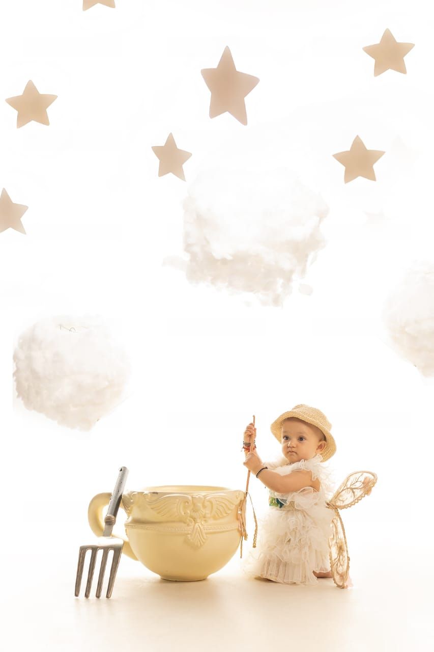 Baby in white dress and hat with fairy wings poses beside a golden teacup amid stars and clouds.
