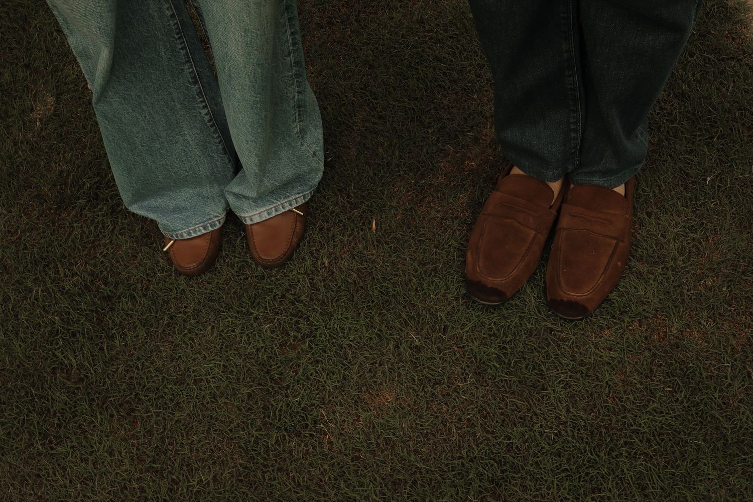Two people standing on grass wearing brown loafers, one in blue jeans and one in gray trousers.