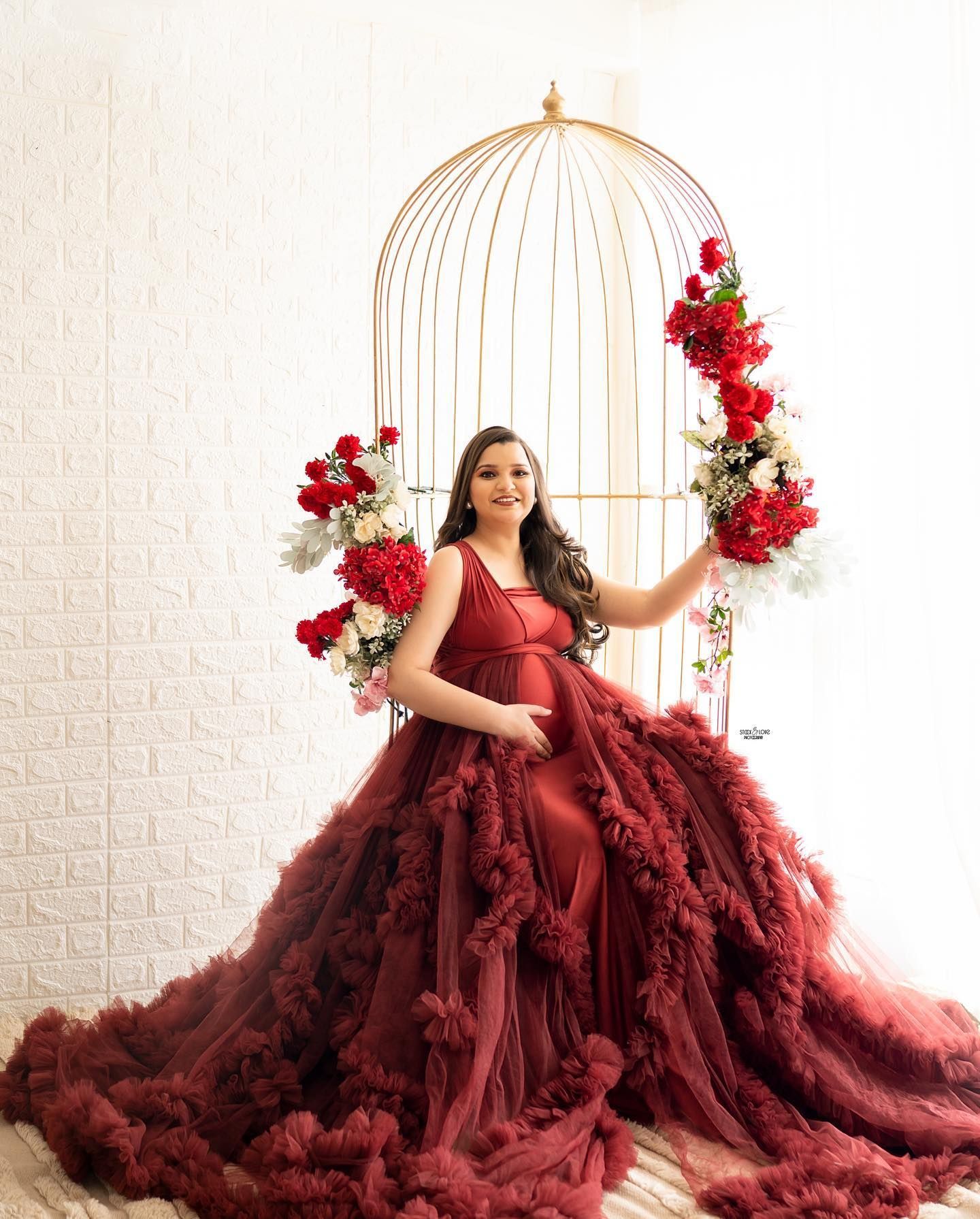 Dramatic maternity portrait with burgundy gown inside golden birdcage frame with red flowers.