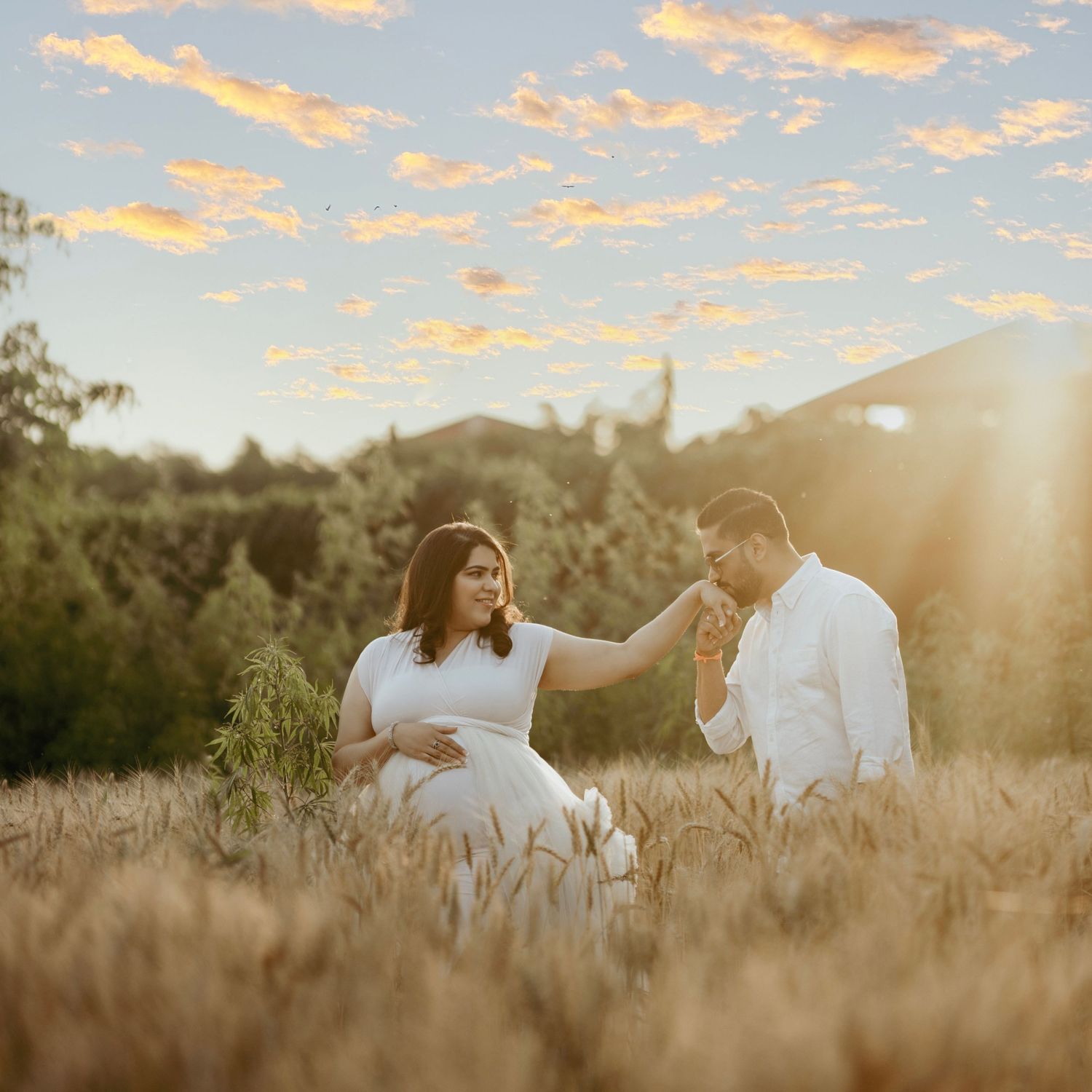 Golden hour maternity session with couple in white outfits walking through wheat field.