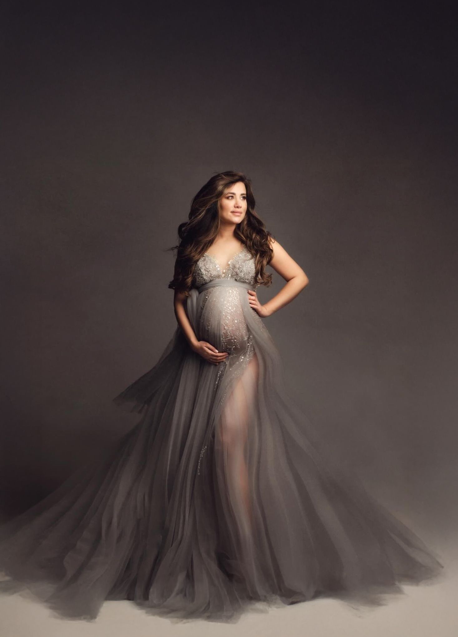 Ethereal maternity portrait in flowing grey tulle gown against dark backdrop.