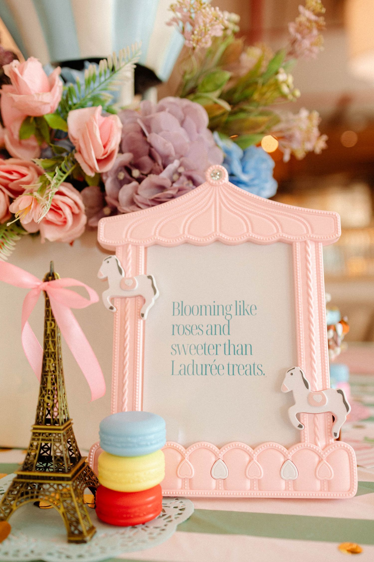 Pink carousel photo frame with floral bouquet, Eiffel Tower figurine, and macarons on a decorated table.