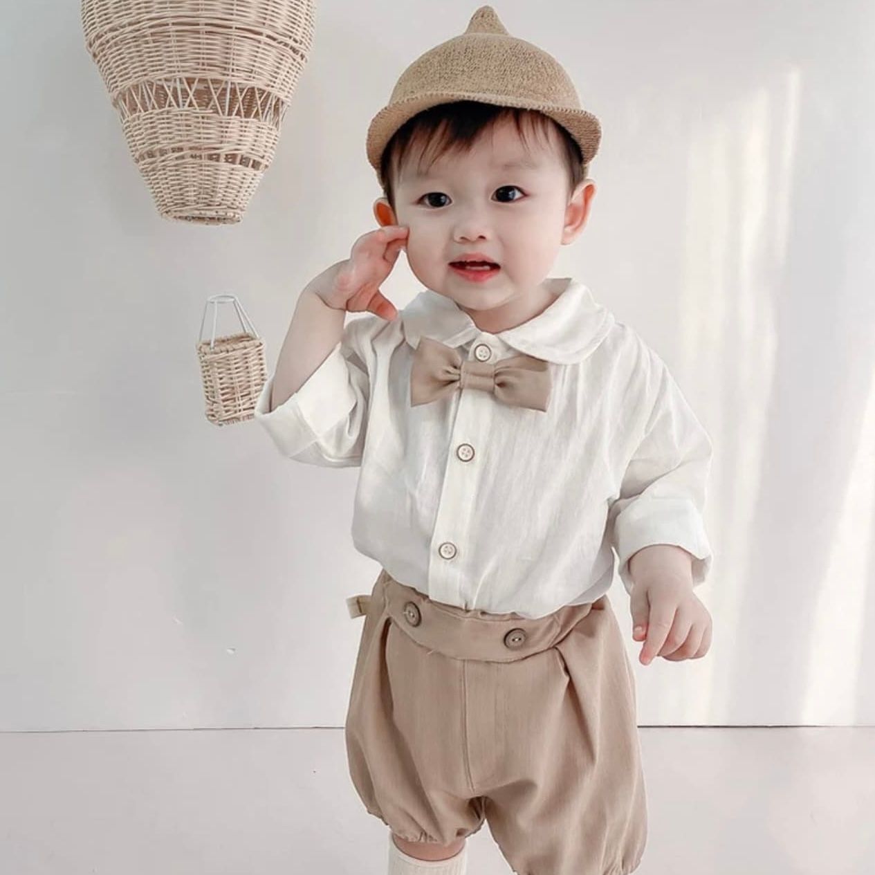 Baby wearing vintage-inspired white shirt and beige romper outfit with suspenders staged against white background.