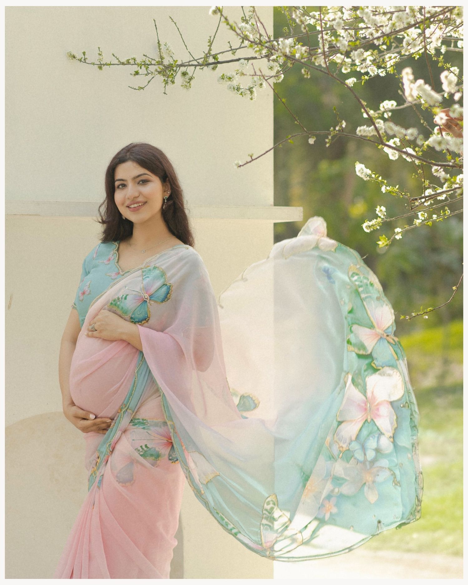 Pregnant woman in pink and teal floral saree poses gracefully outdoors near blooming white flowers.