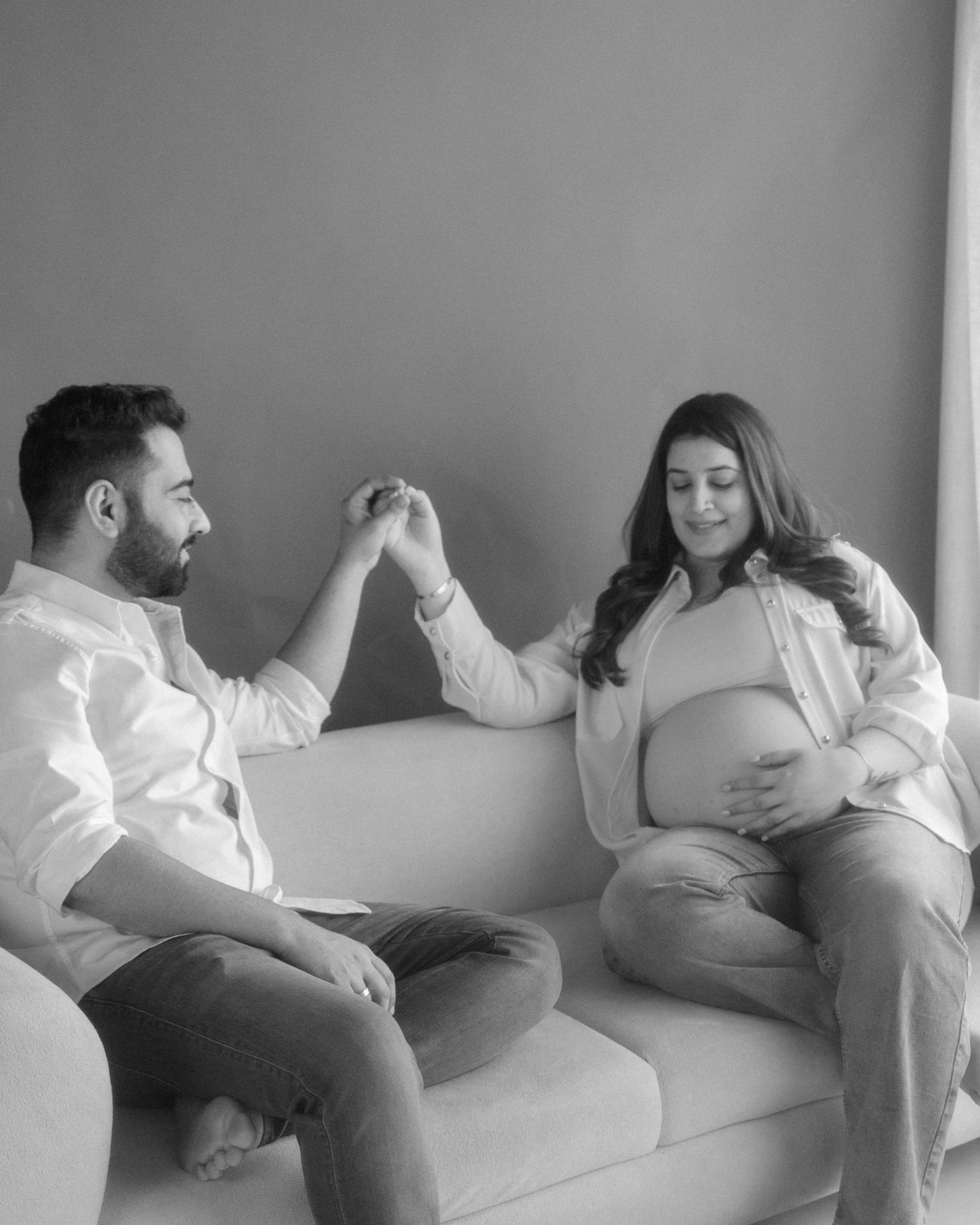 Black and white maternity photo of couple holding hands on sofa, pregnant woman cradling baby bump.