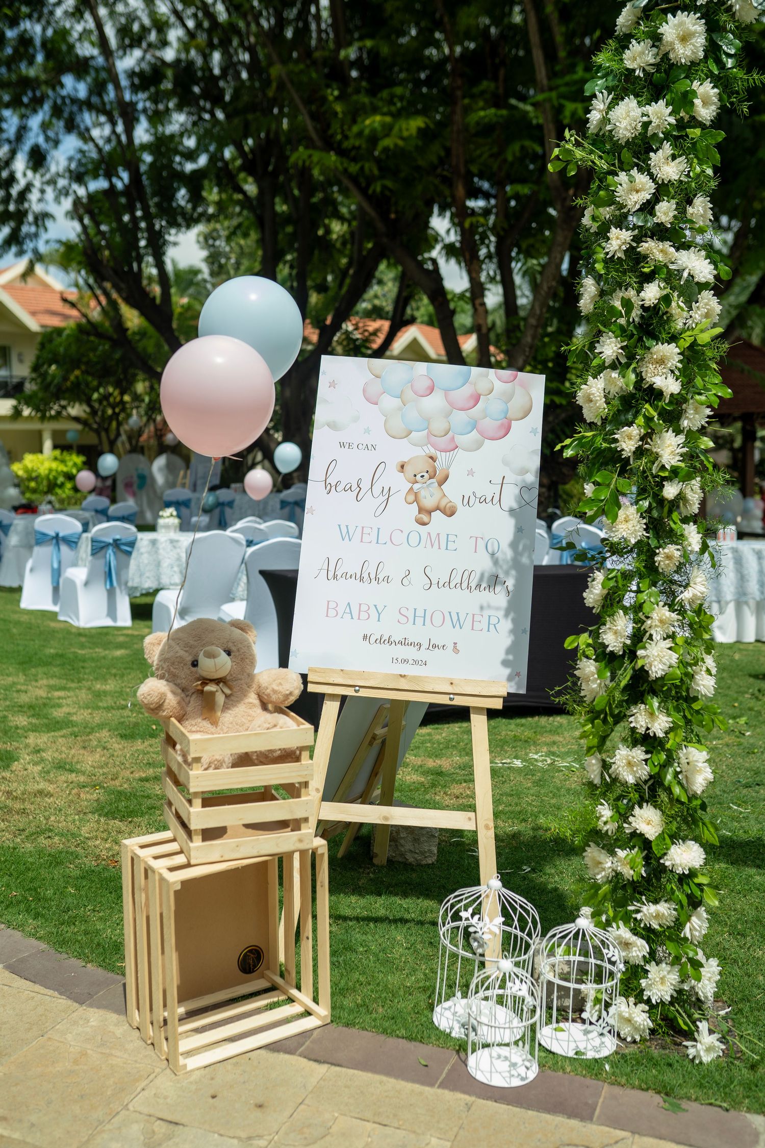 Outdoor baby shower welcome sign on easel with teddy bear, pink and blue balloons, wooden crates, and floral arch decoration.