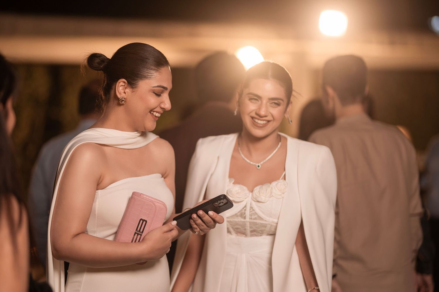 Two women in white outfits smiling at a phone at an elegant evening event.