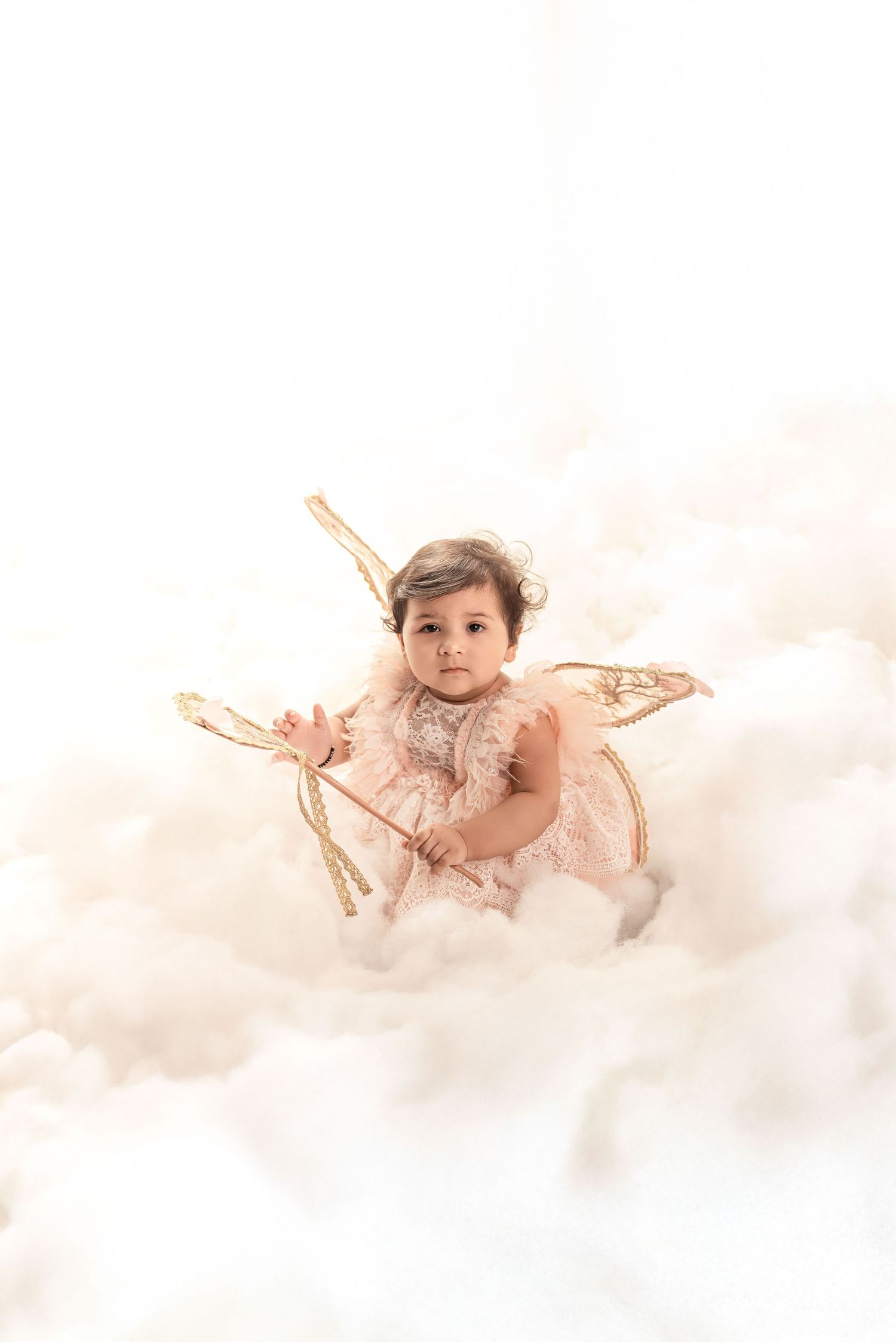 Adorable baby dressed as an angel with gold wings and wand sitting among fluffy white clouds.