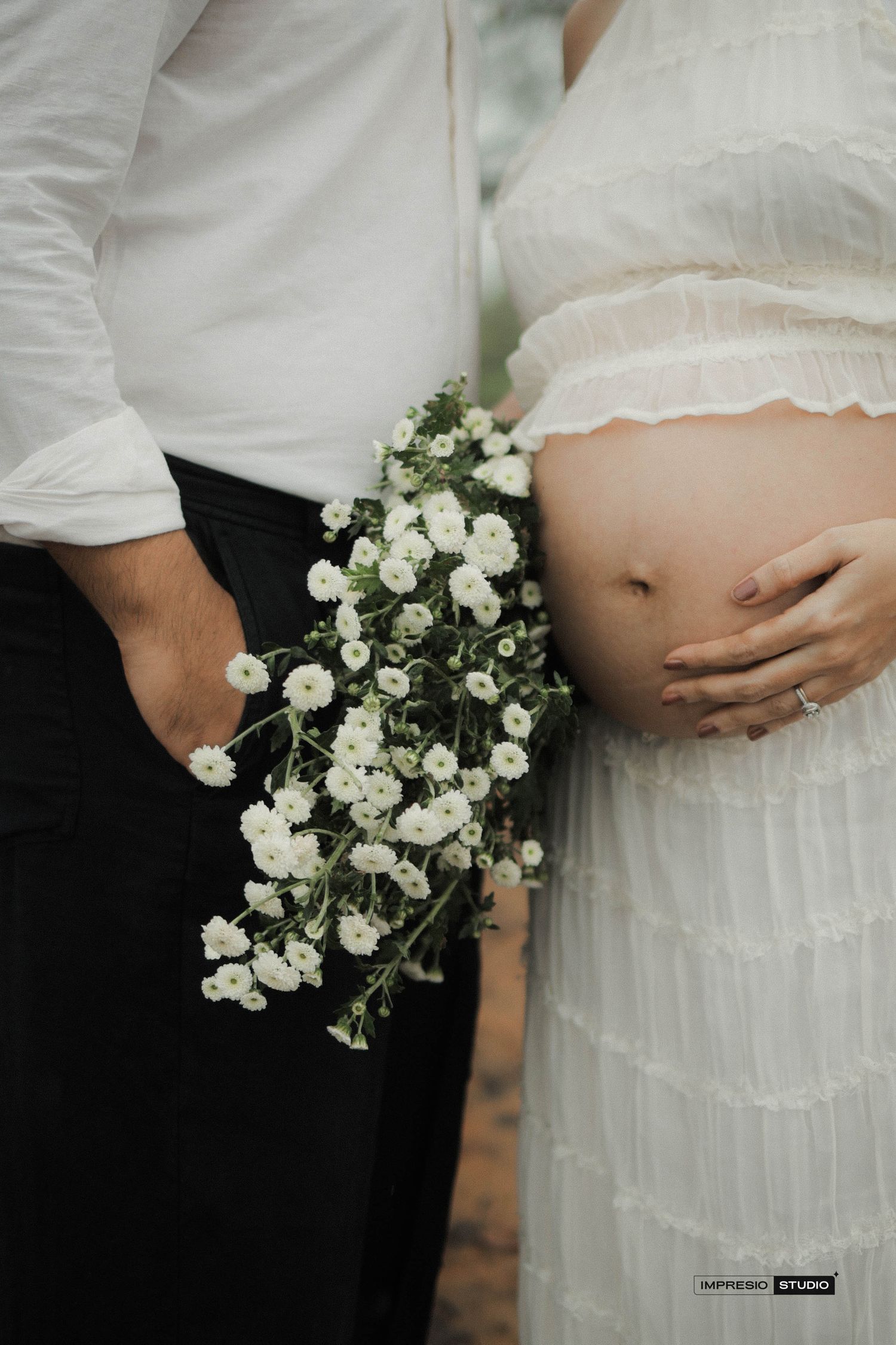 Couple maternity photo with pregnant belly, white flowers bouquet, white dress, and black pants by Happiness Studio.