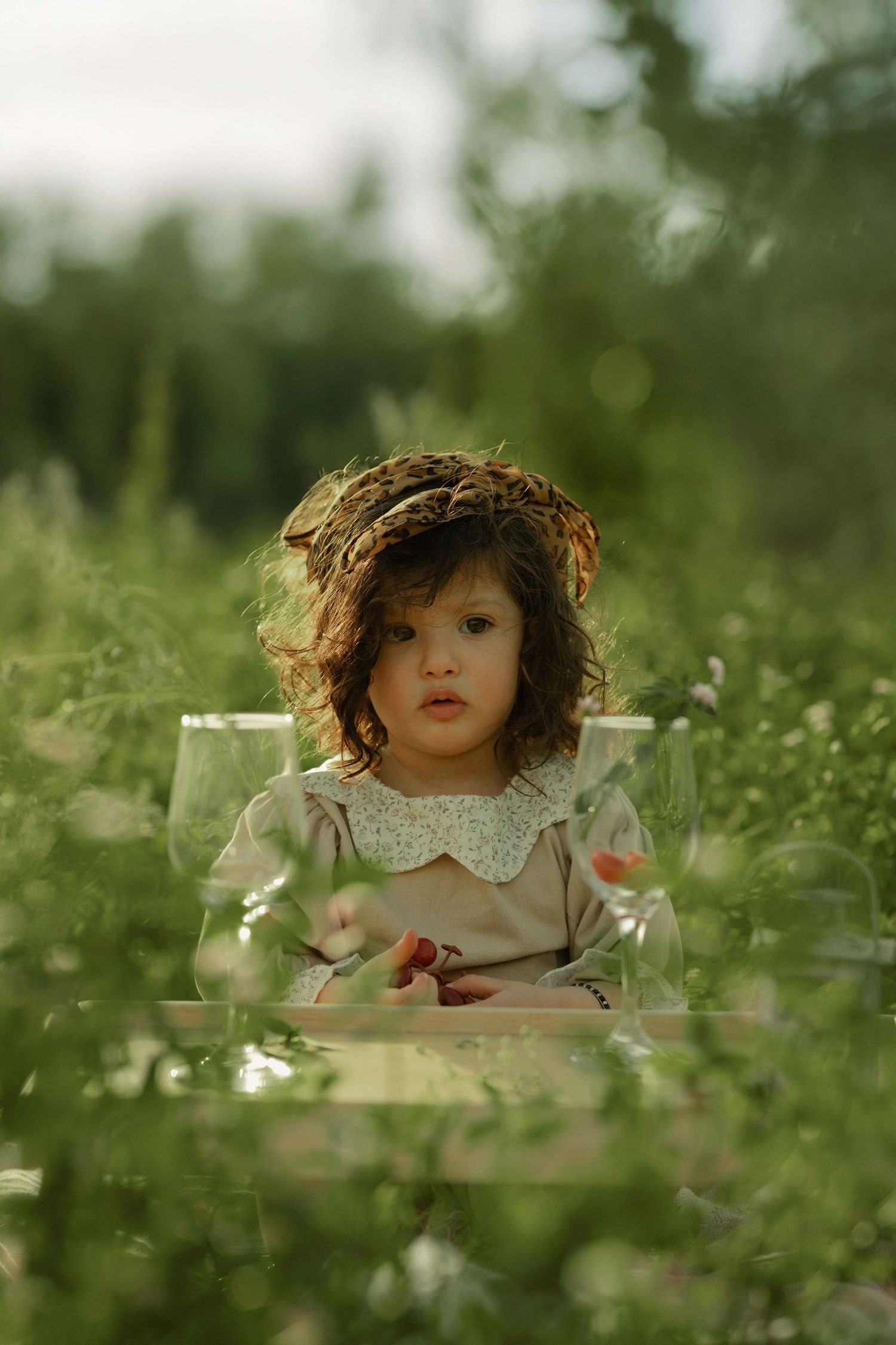 A young girl with curly hair and a floral wreath sits among lush green grass wearing a vintage-style dress.