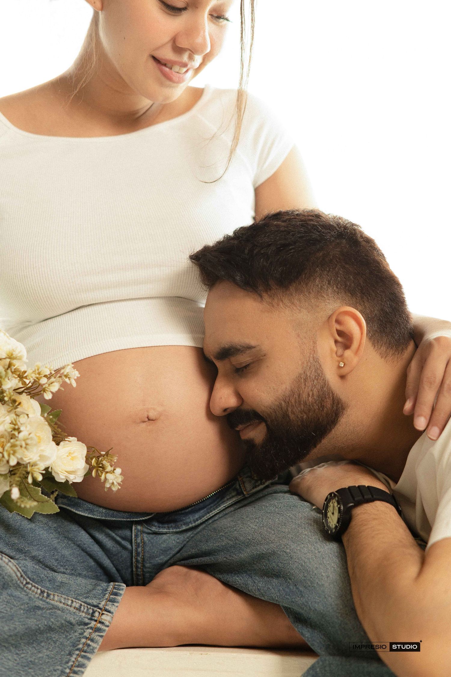 A man tenderly rests his head on a pregnant woman's belly during a maternity photo shoot with white flowers.