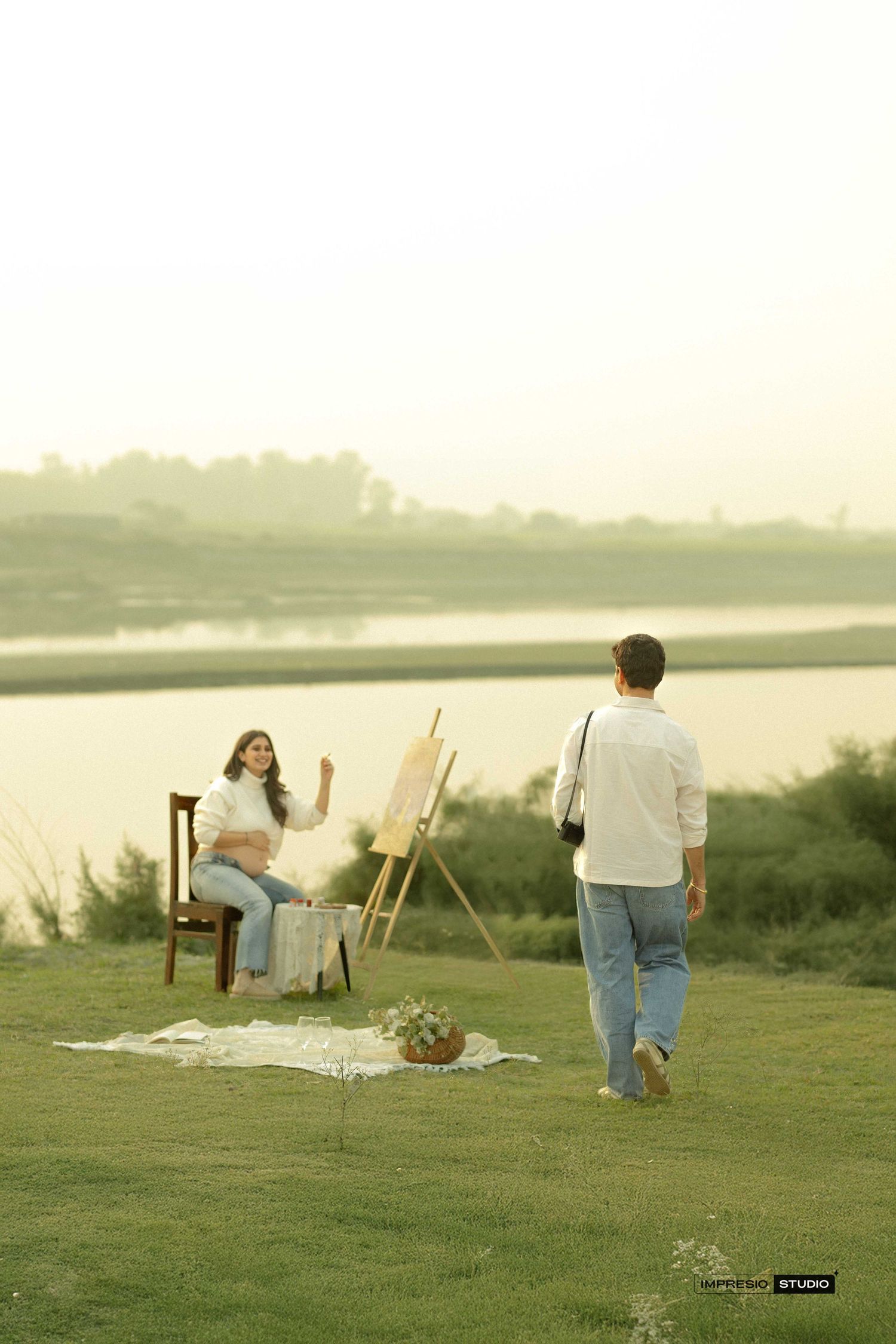 Pregnant woman sitting on chair near easel by lake at sunset, man with camera approaching during outdoor maternity photoshoot.