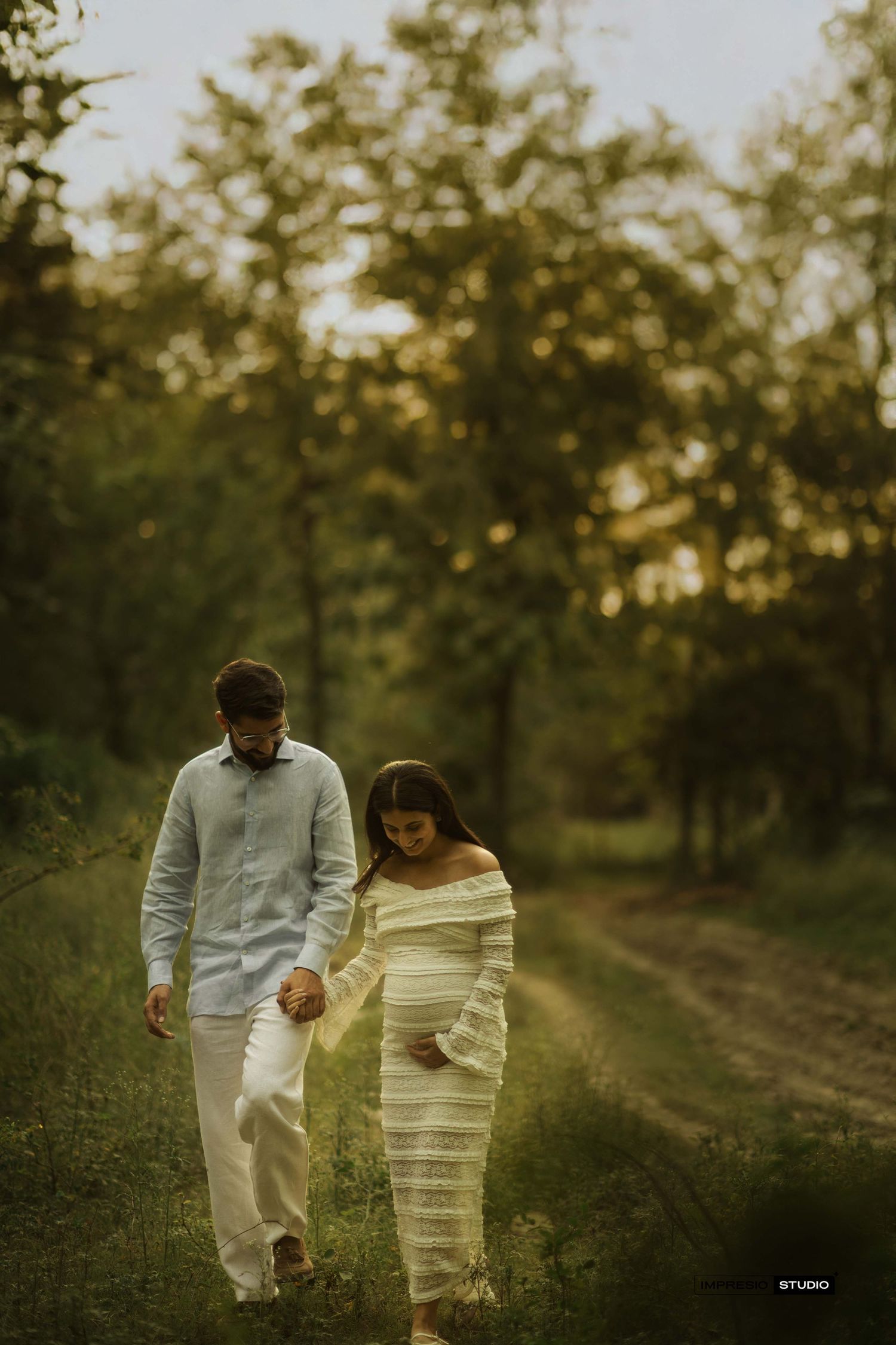 Couple walking outdoors during golden hour maternity photoshoot, woman in striped off-shoulder dress cradling baby bump.