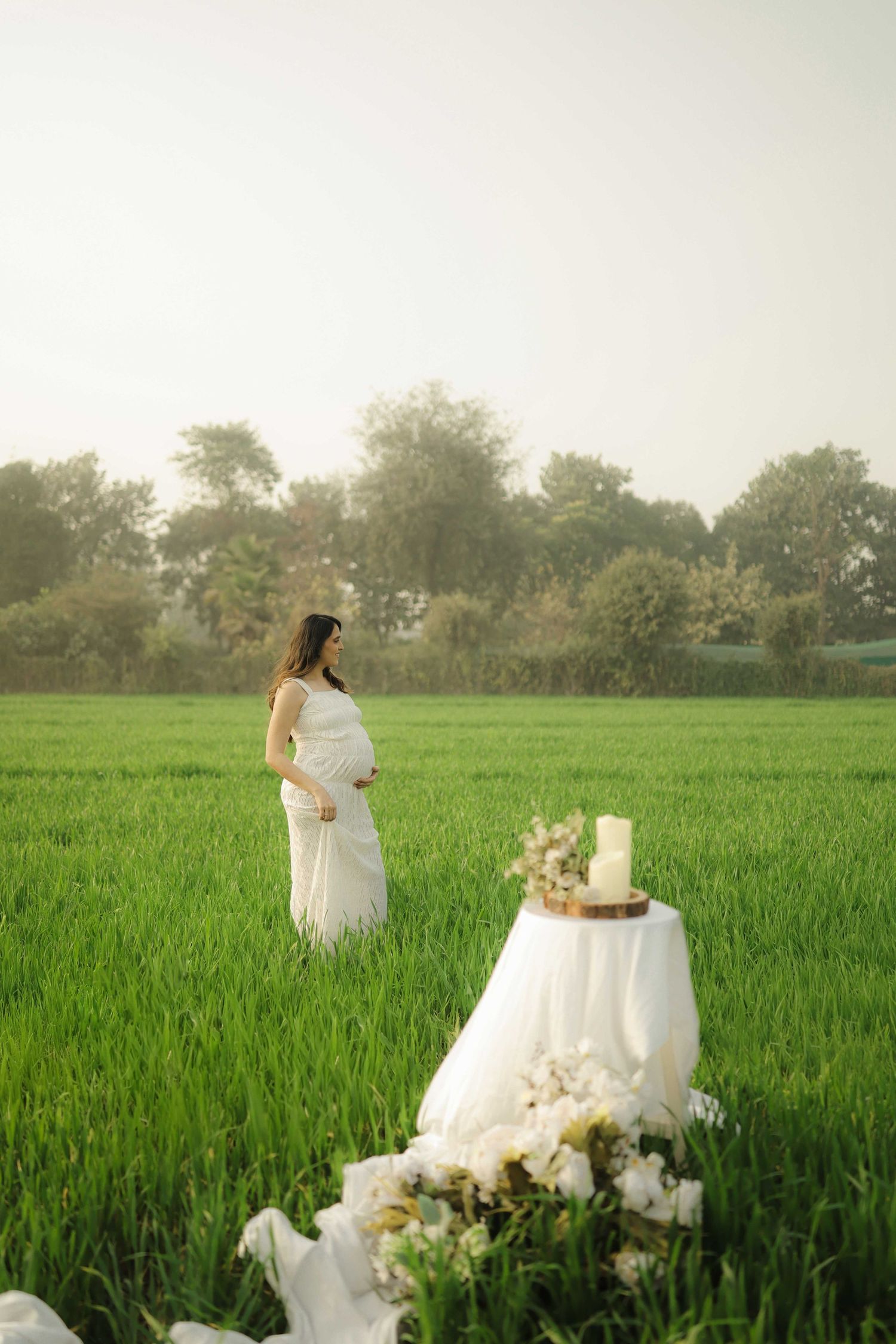 Pregnant woman in white dress standing in lush green field beside a decorated table with cake and flowers.