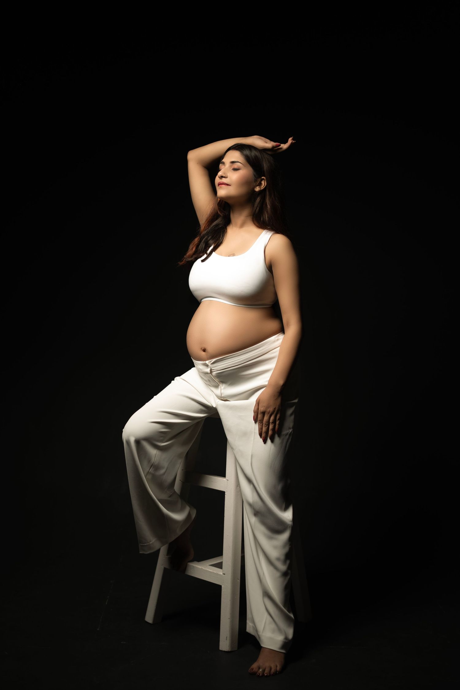 Pregnant woman in white crop top and pants posing gracefully on stool against black background.