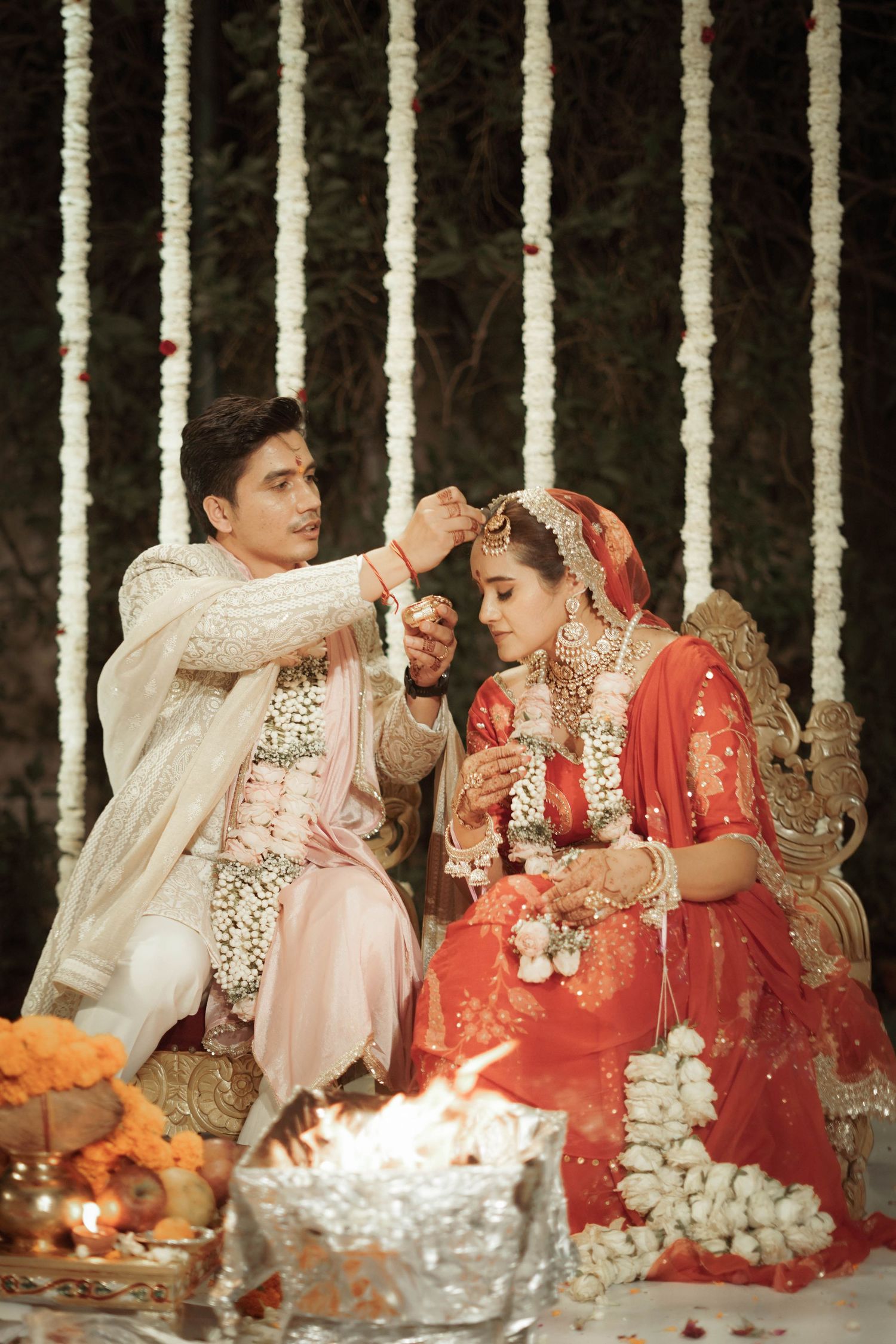 Groom applying sindoor to bride in red lehenga during traditional Hindu wedding ceremony with floral backdrop.