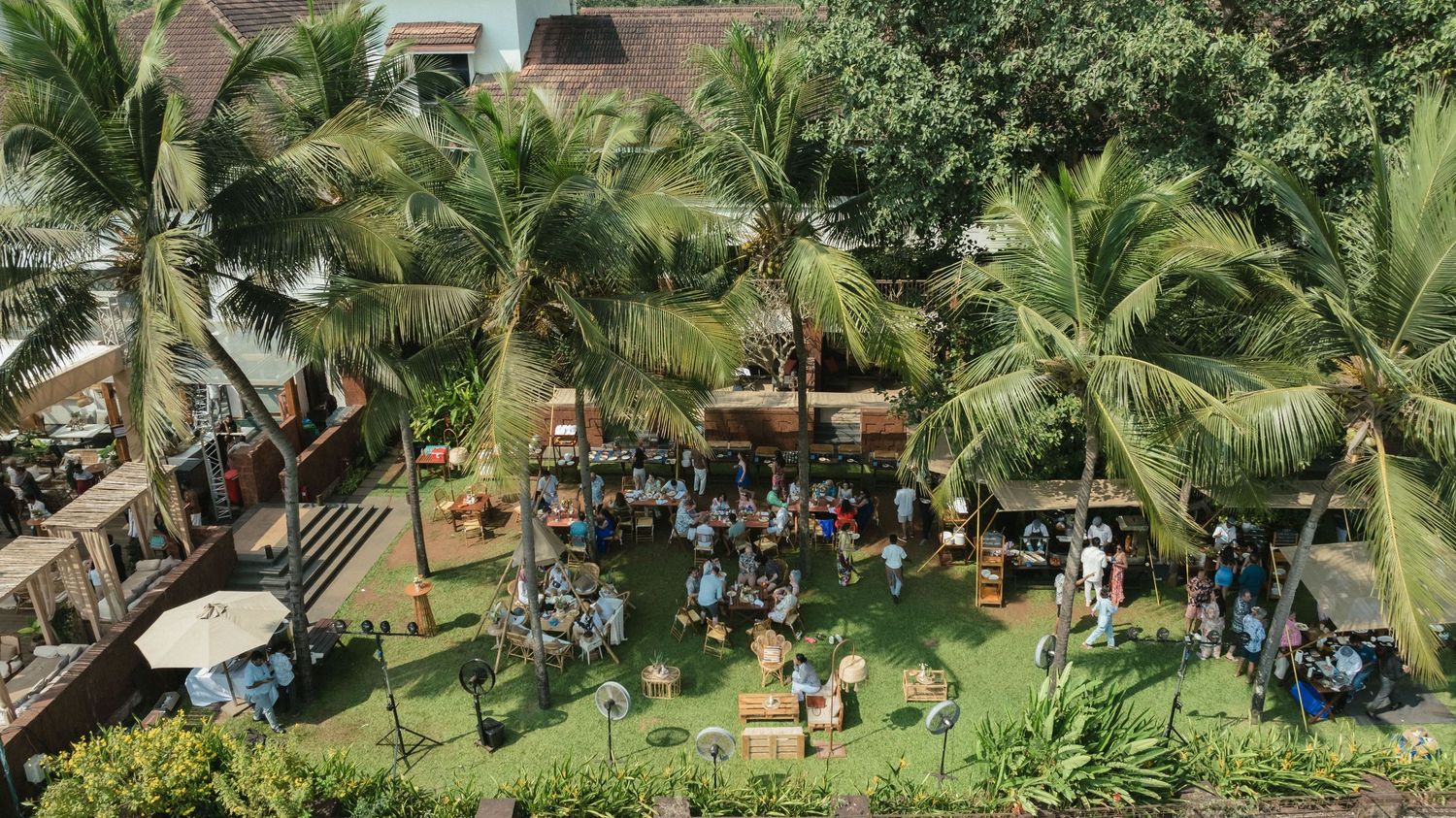 Aerial view of a tropical outdoor market event with crowds gathered among tall palm trees on a lush green lawn.
