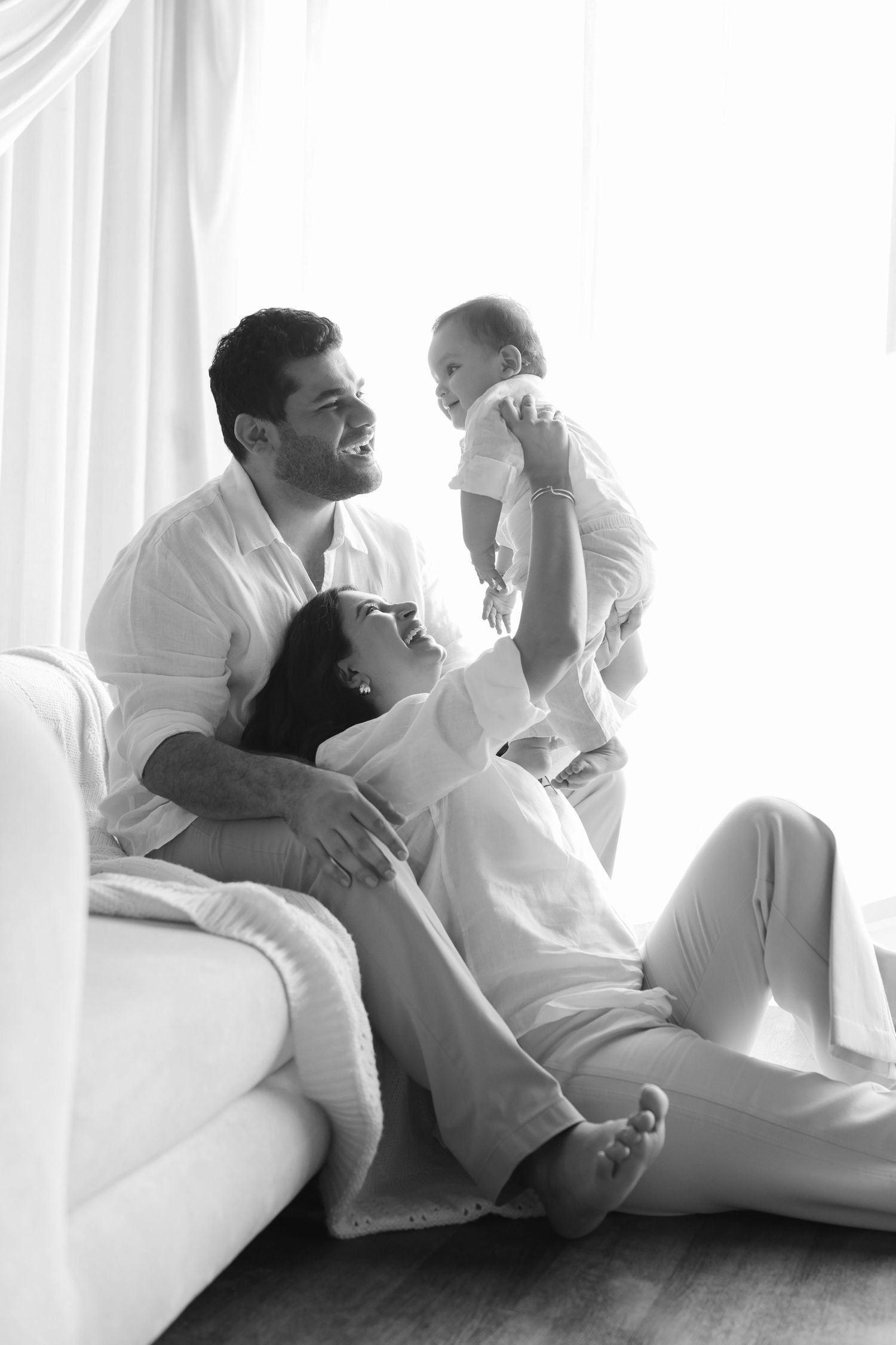 Black and white photo of happy family with mother, father, and baby sitting together on floor near bright window.
