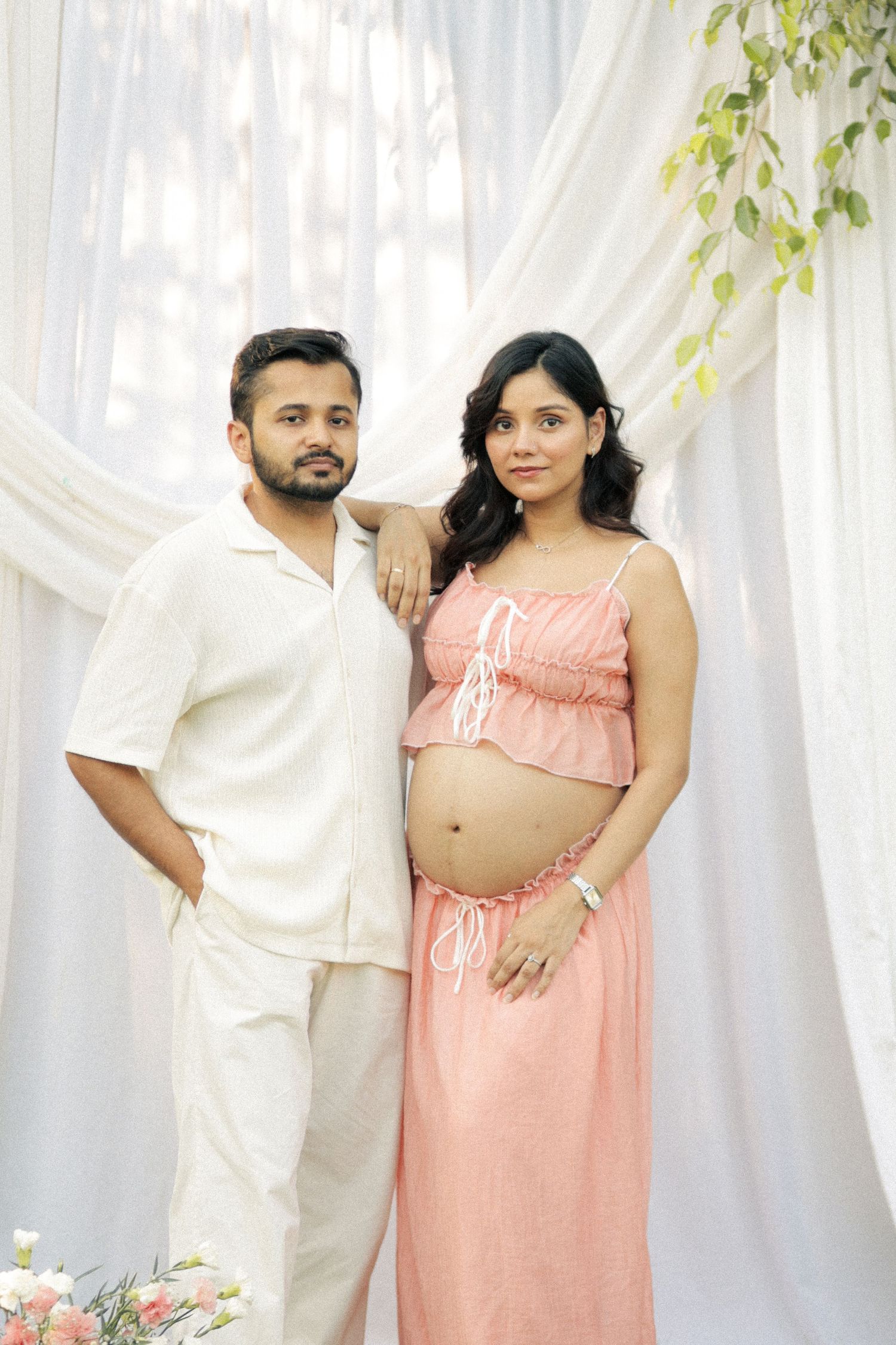 Pregnant couple posing together, man in white outfit, woman in pink maternity dress, white drape backdrop.