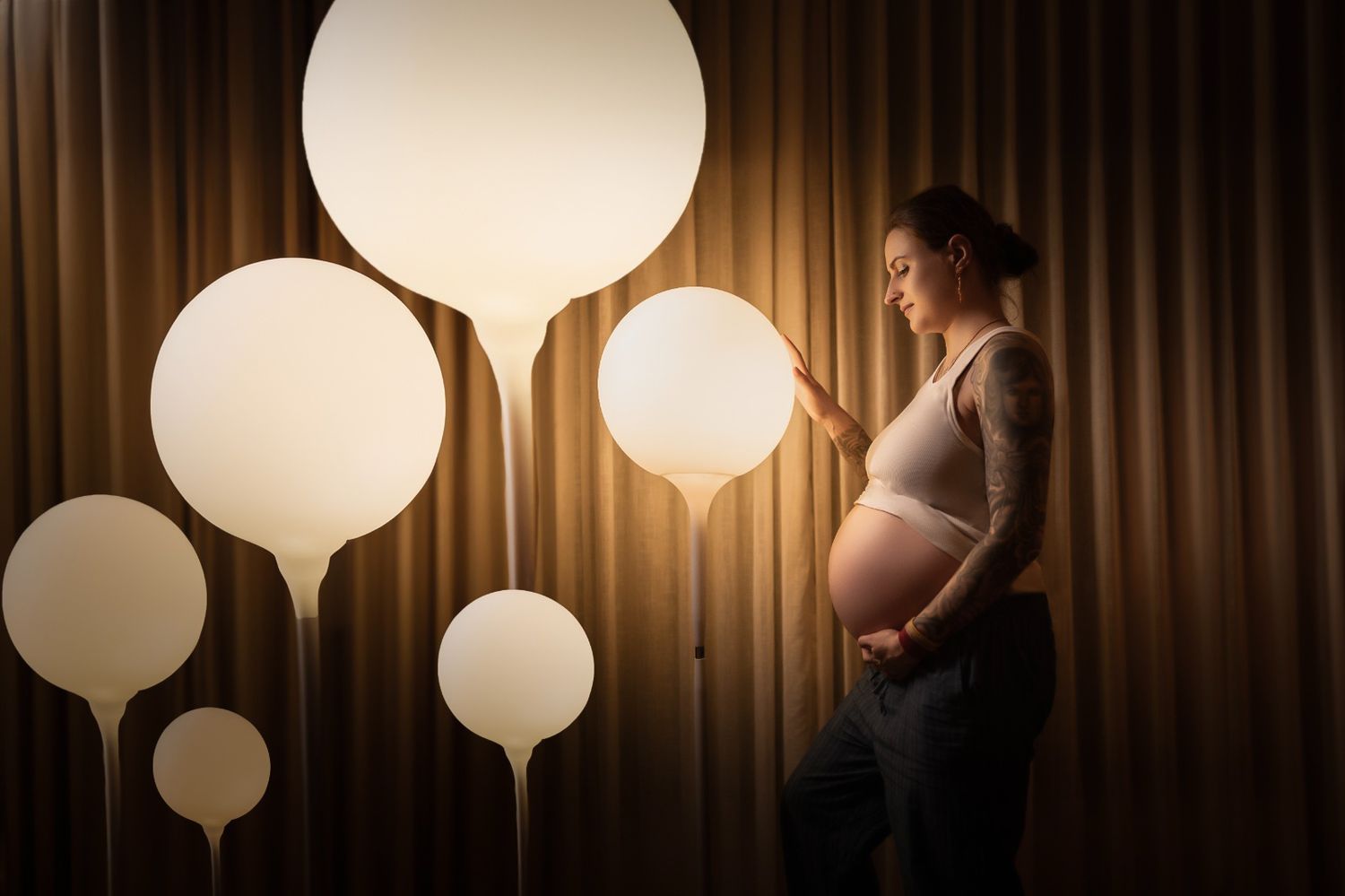 A pregnant woman stands beside glowing white balloon lights against a brown curtain backdrop in dramatic lighting.