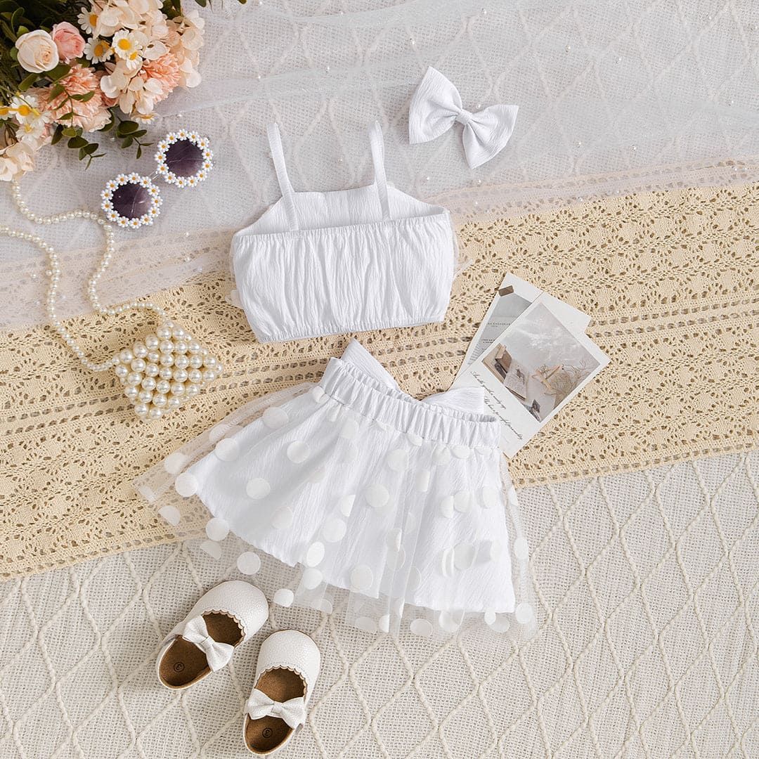 White baby girl outfit with crop top, tulle skirt, bow accessory, and brown shoes laid on textured fabric backdrop.
