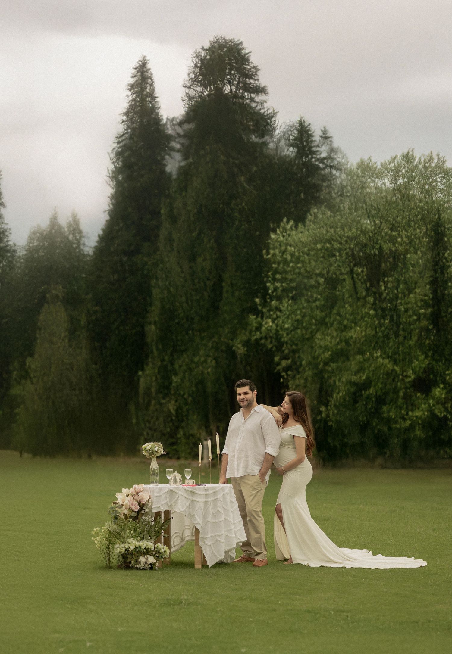 Couple in white attire standing by a floral decorated table on a lush green lawn with tall evergreen trees.