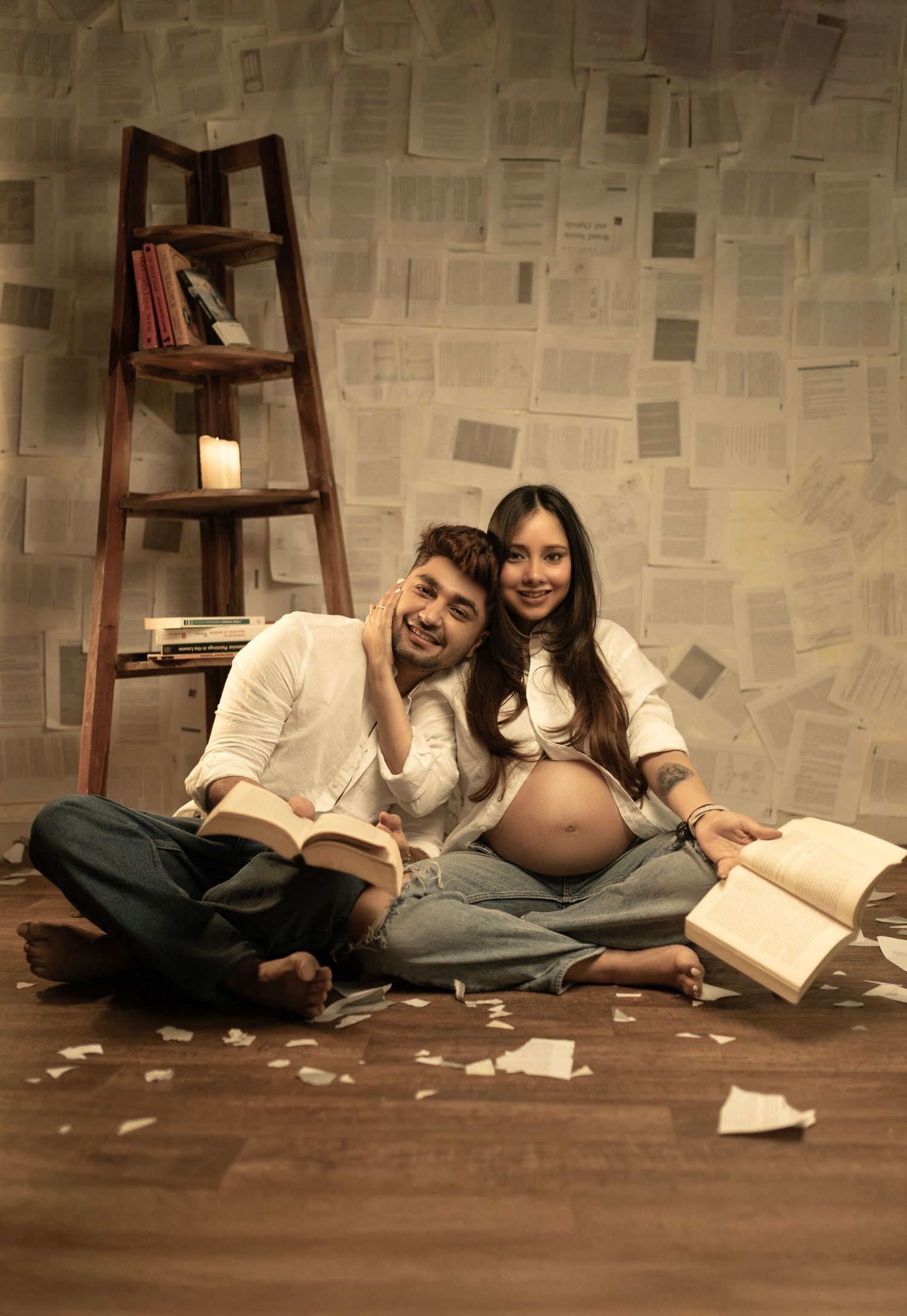 Smiling couple sitting on floor with books, pregnant woman in white top, ladder bookshelf, pages on wall backdrop.