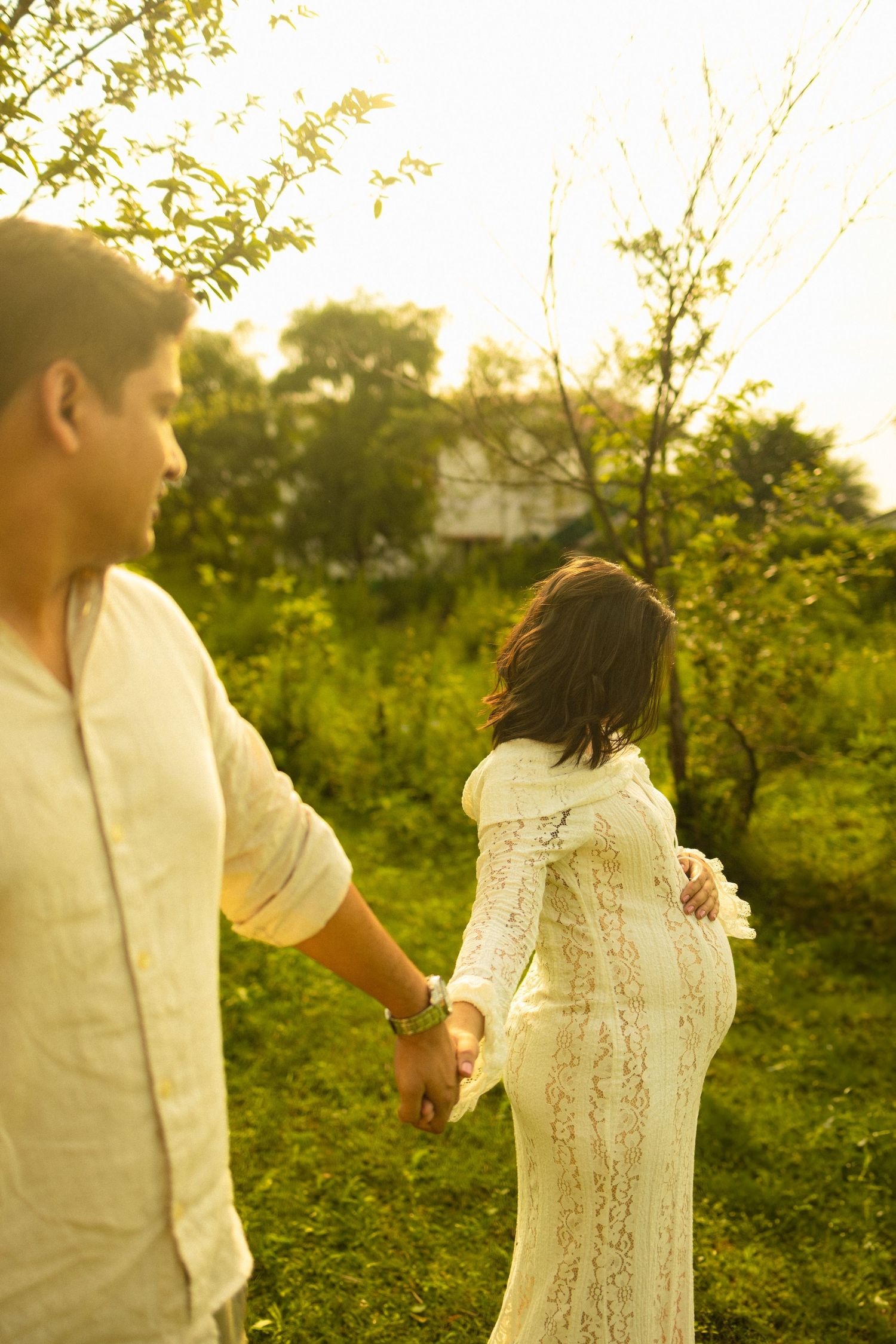 A couple holds hands while walking through a lush green garden in golden sunlight, the woman wearing a white lace dress.