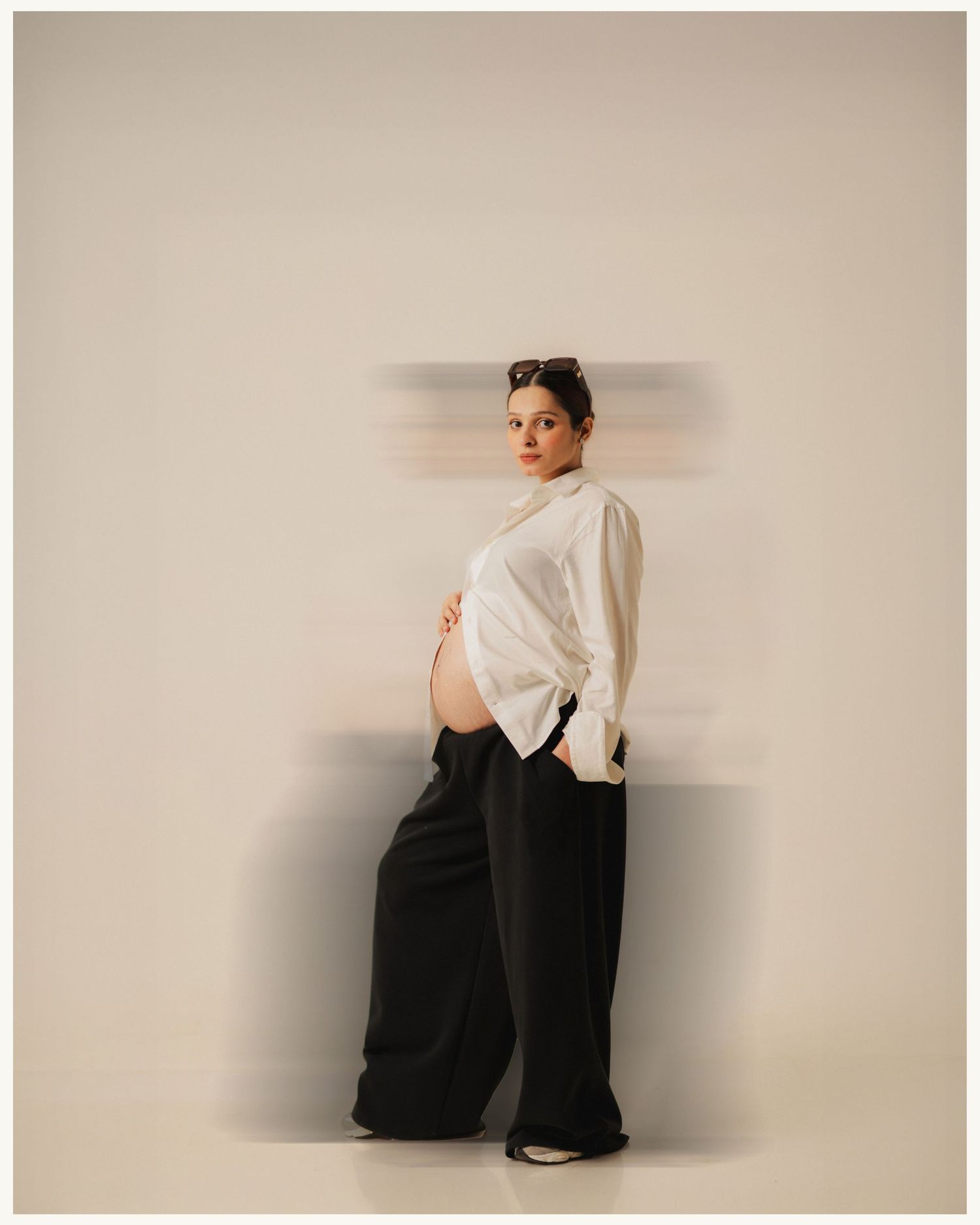Pregnant woman in white blouse and black wide-leg pants poses gracefully with motion blur effect on neutral background.