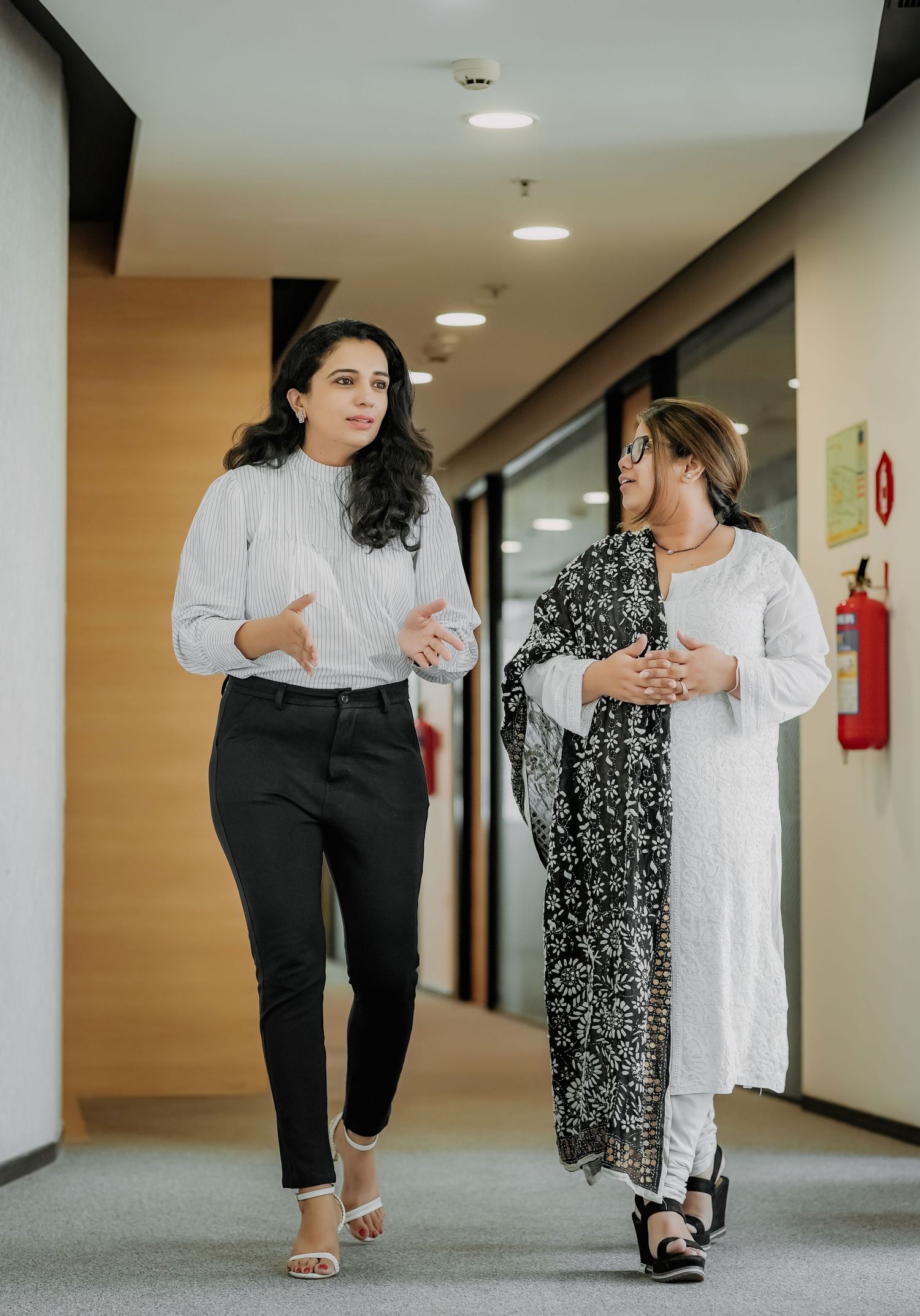 Two professional women walking and talking in a modern office corridor, one in white blouse and black pants.