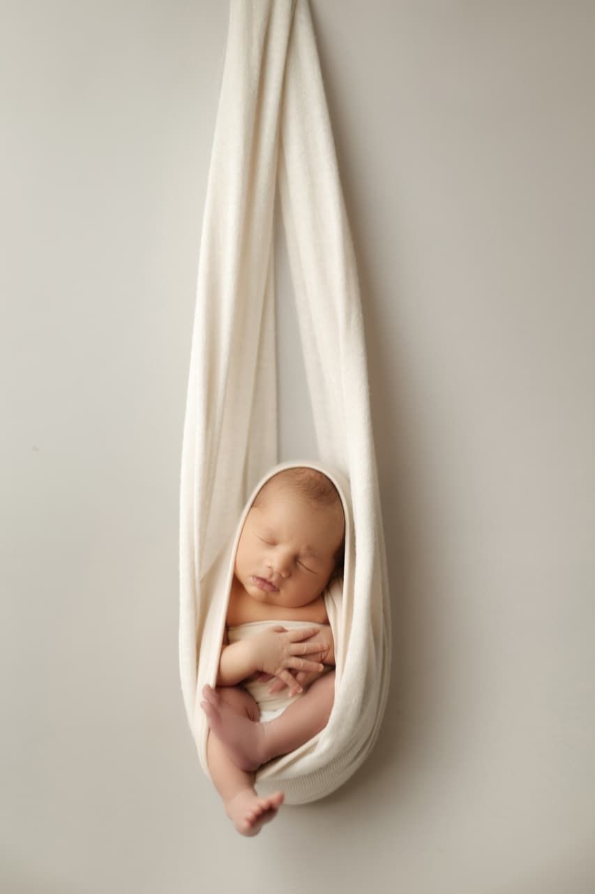 Newborn baby sleeping peacefully in a cream fabric sling against a soft white background.