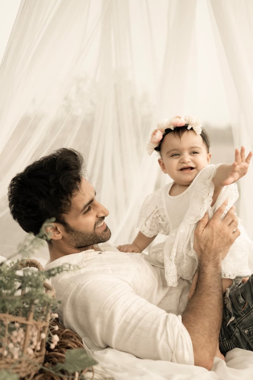 A father lovingly holds his smiling baby girl wearing a floral headband in a soft, dreamy white setting.