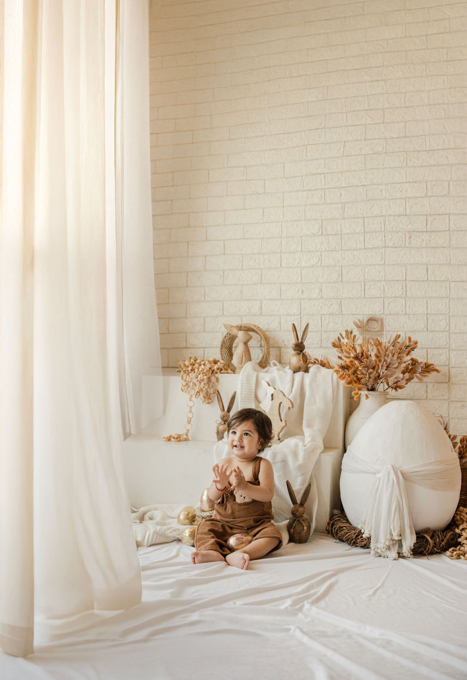 A baby in brown overalls sits on white fabric near Easter decorations and dried flowers against a cream brick wall.
