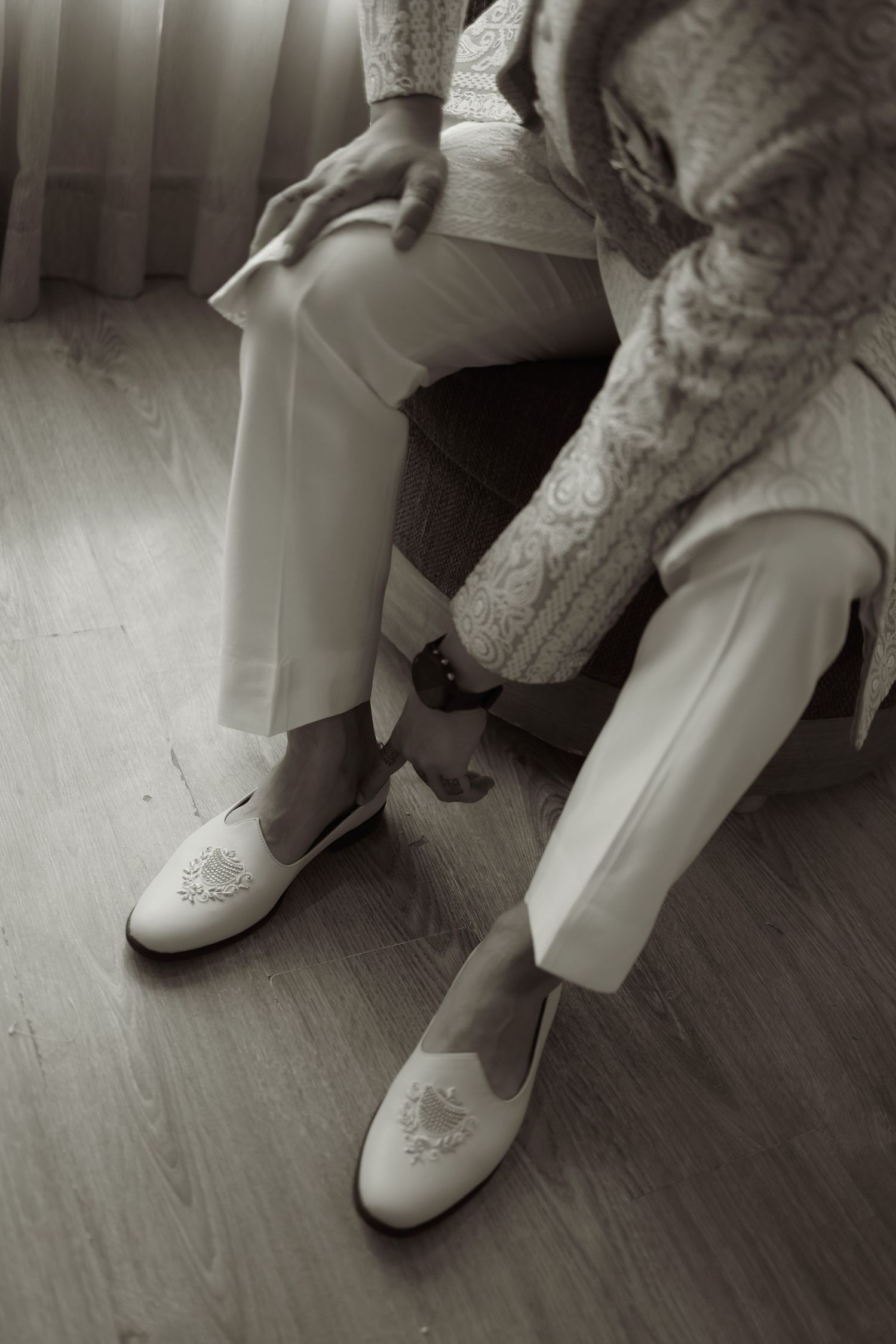 Close-up of groom wearing white embroidered loafers and white trousers while sitting in a chair on wedding day.