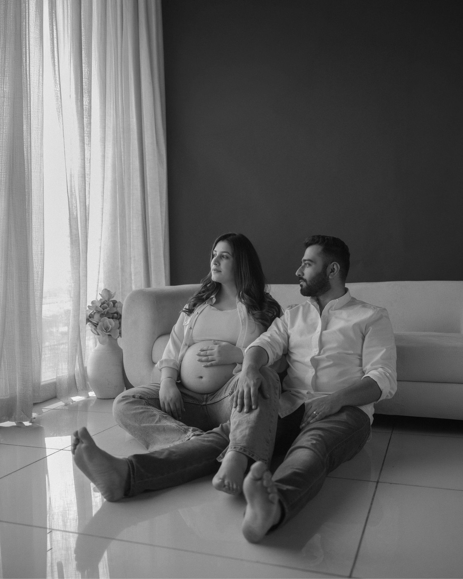 Black and white photo of a pregnant couple sitting on floor leaning against a sofa near a bright window.