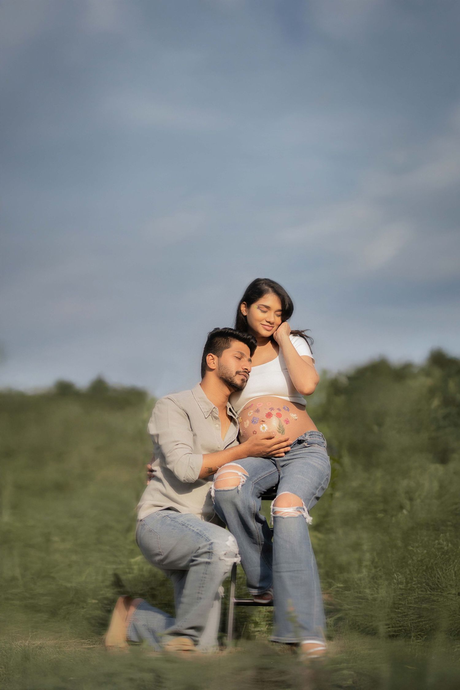 A couple poses outdoors, woman with painted pregnant belly sitting while man kneels beside her in a green field.