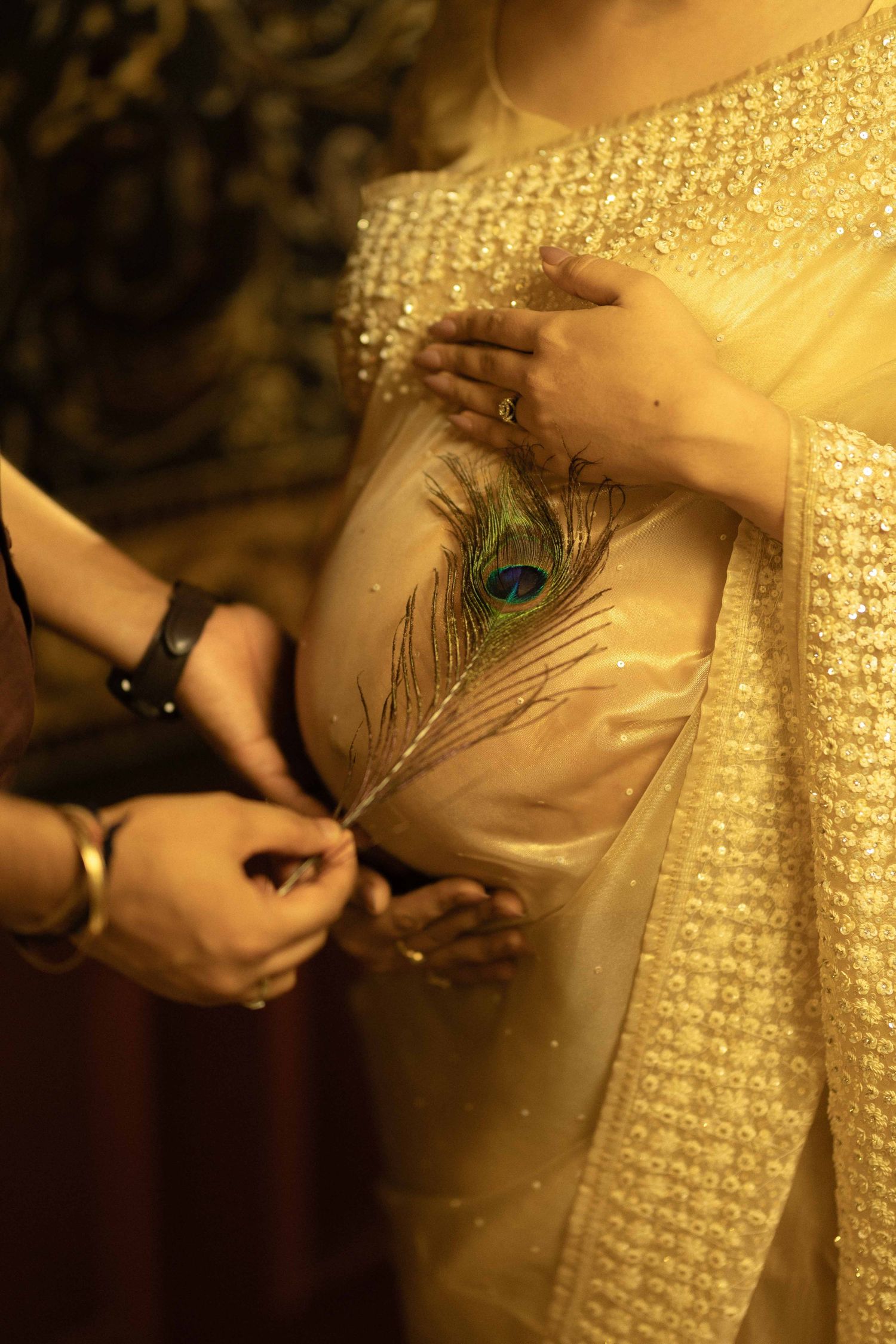 Hands holding a peacock feather against a cream embroidered saree during an Indian wedding ceremony.