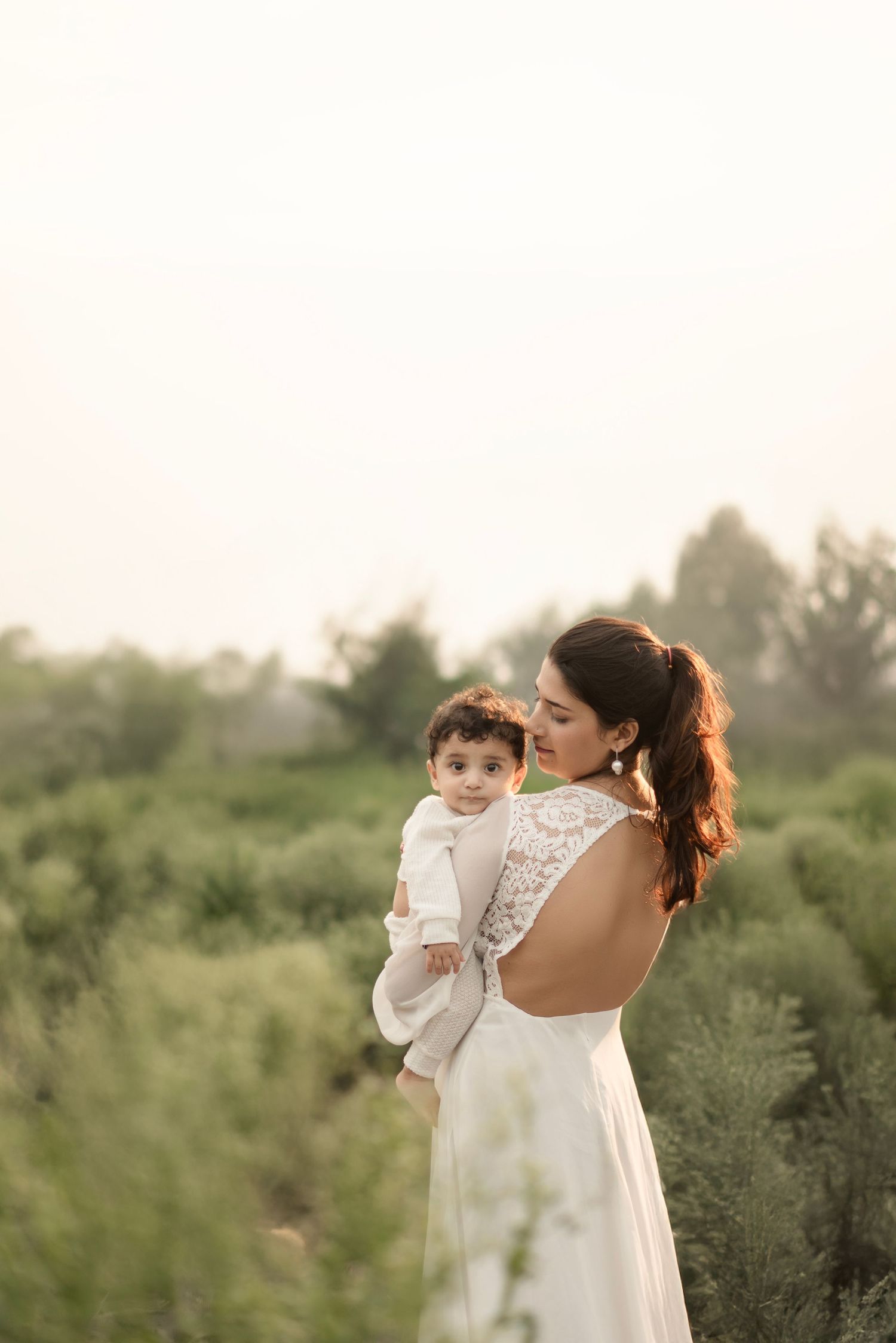 A bride in a white lace-back gown holds a baby in an outdoor setting with lush green trees.
