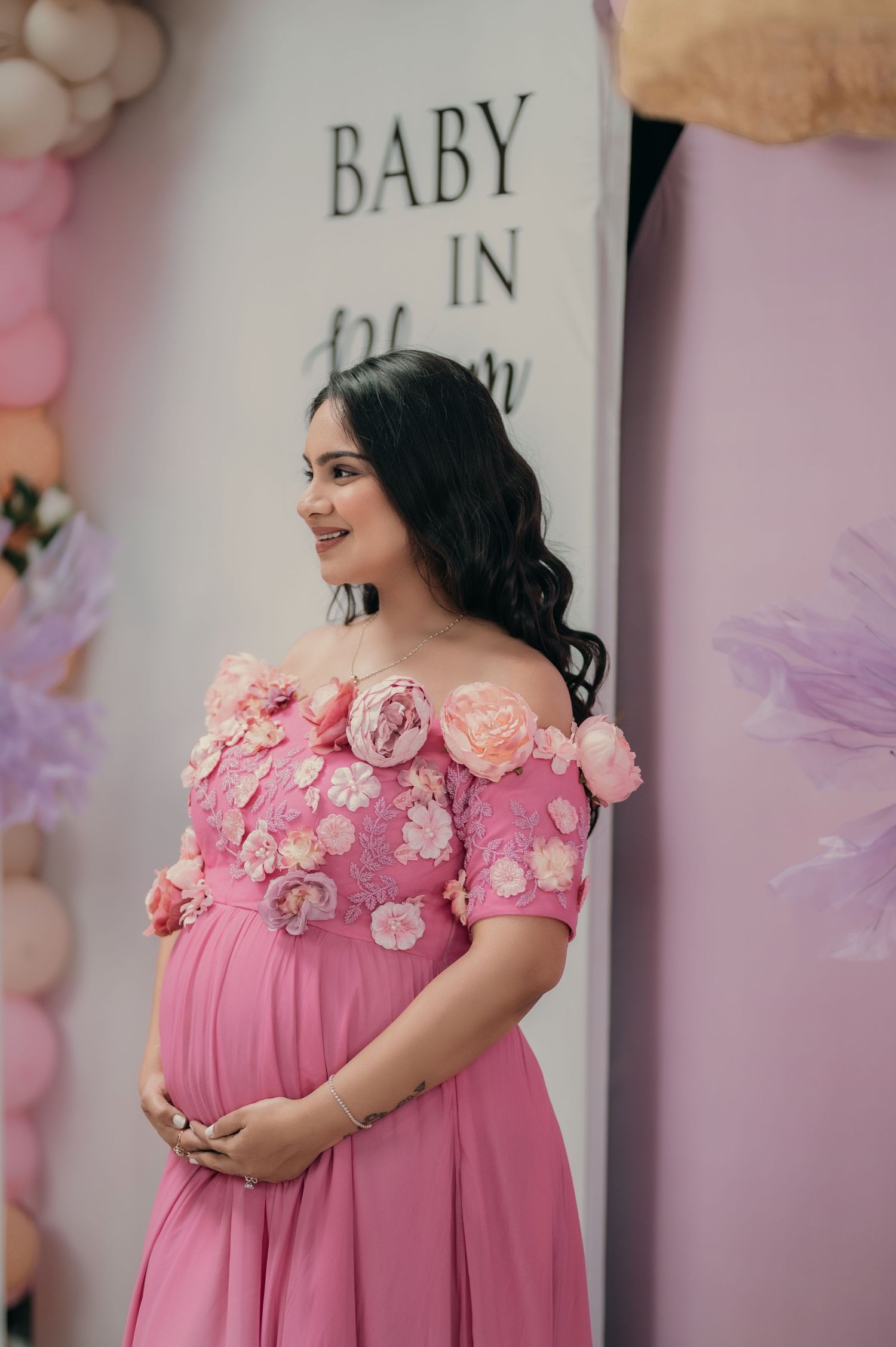 Pregnant woman in pink floral off-shoulder gown smiling at baby shower with balloon decorations.