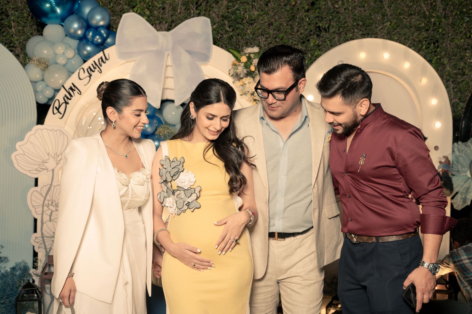 Pregnant woman in yellow dress at baby shower with three guests posing in front of balloon decorations.
